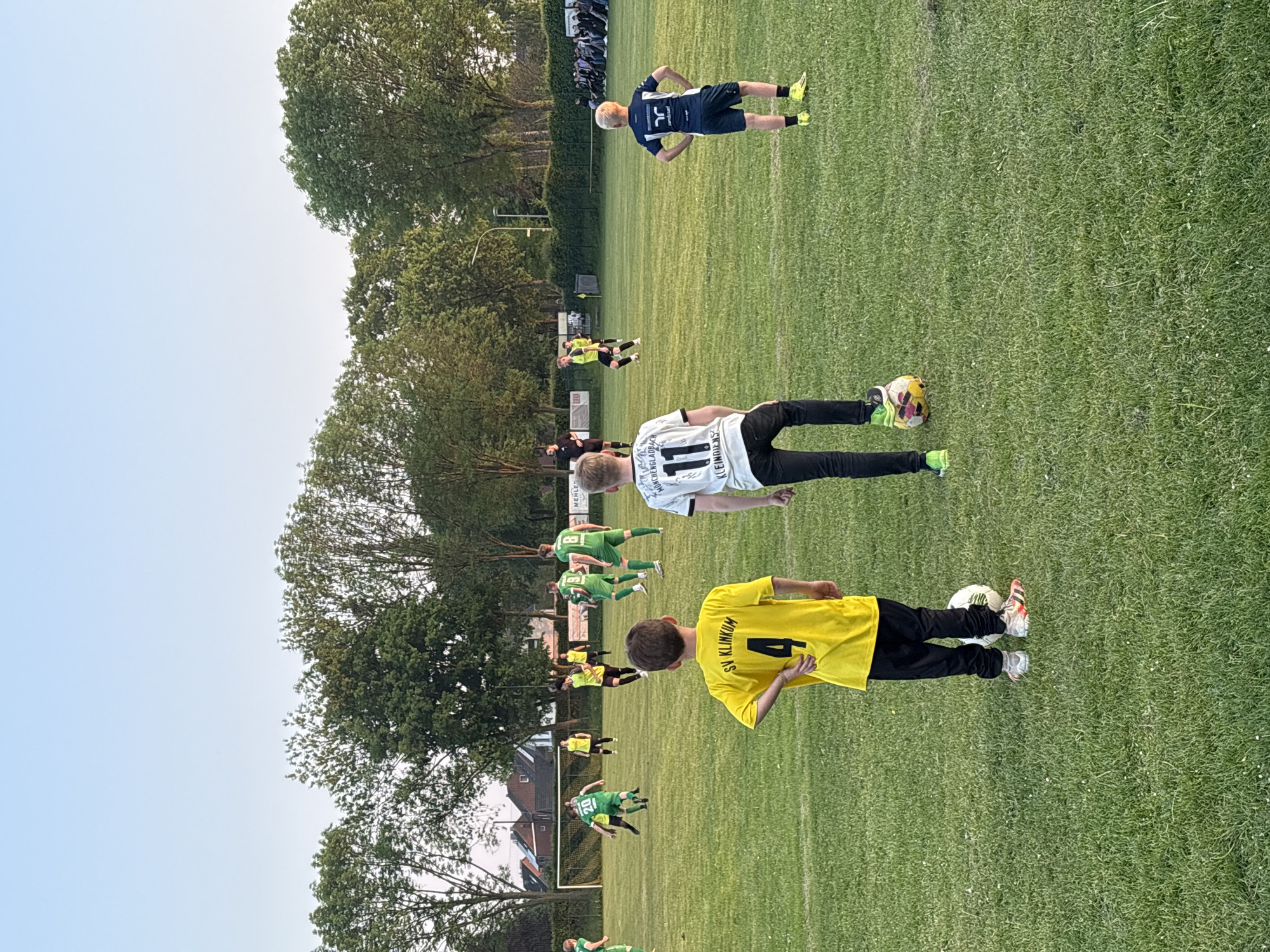 The image depicts a soccer game in Wegberg, Germany. Several players in green and yellow uniforms are visible on a green field, along with at least two players in different uniforms (one in white, one in dark blue). One player is wearing a yellow jersey with the number 4, another a white jersey with the number 11. A tree line is visible in the background, and some buildings are also seen. There are also spectators in the background on the upper right of the picture.