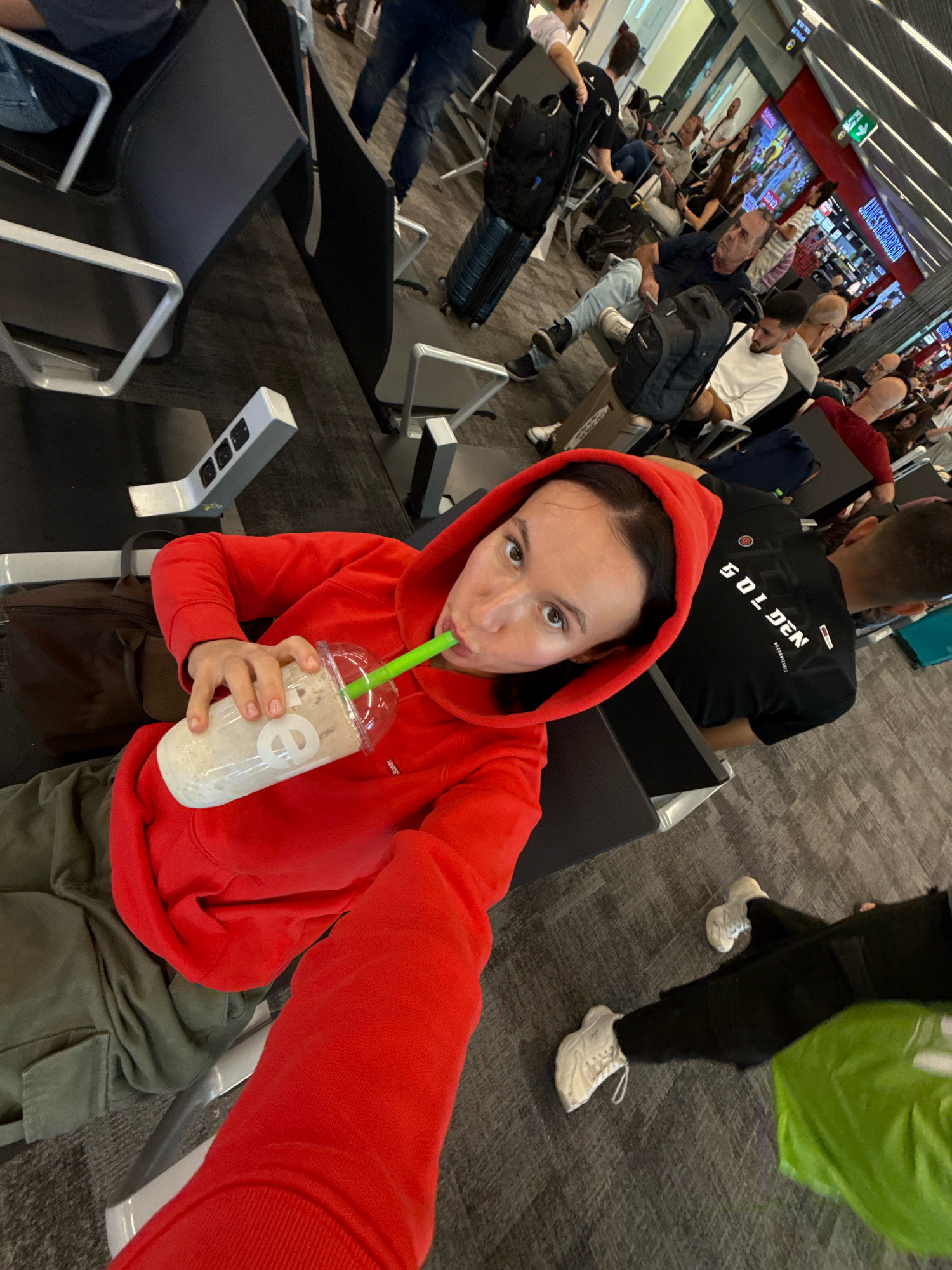 A woman in a red hooded sweatshirt and green pants drinks from a clear plastic cup with a green straw. She is seated in an airport waiting area, with other people visible in the background. The environment appears to be an airport terminal, with rows of seats and luggage. The image is taken from a high-angle perspective, looking down at the woman.