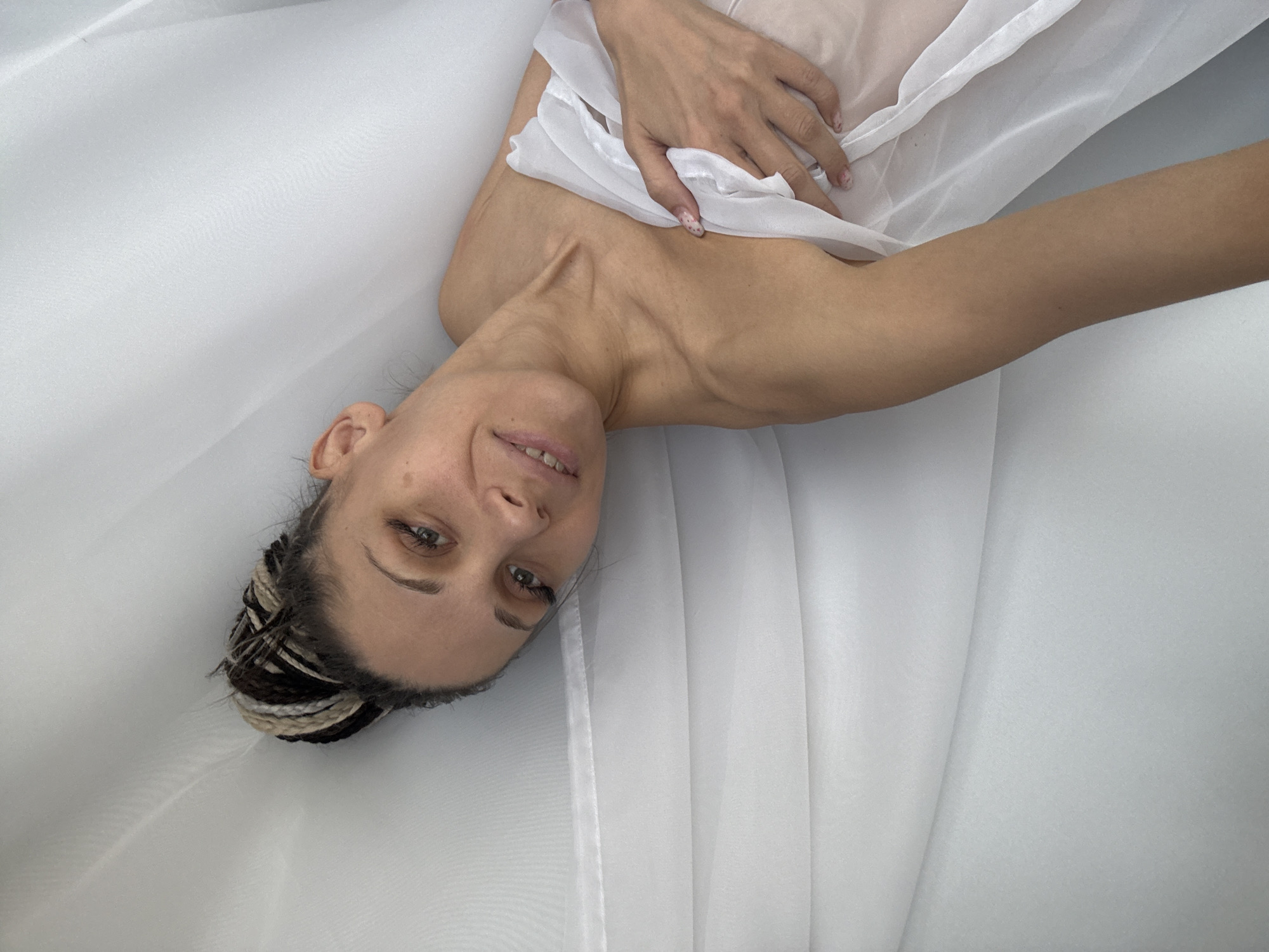 A woman lies in a white bathtub, covered with a sheer white fabric. Her head is tilted to the left, with braided hair. She is smiling slightly. Her left hand is resting on her chest. The scene appears to be indoors. This image does not provide enough information to confirm the location as Rusanovo, Russia.