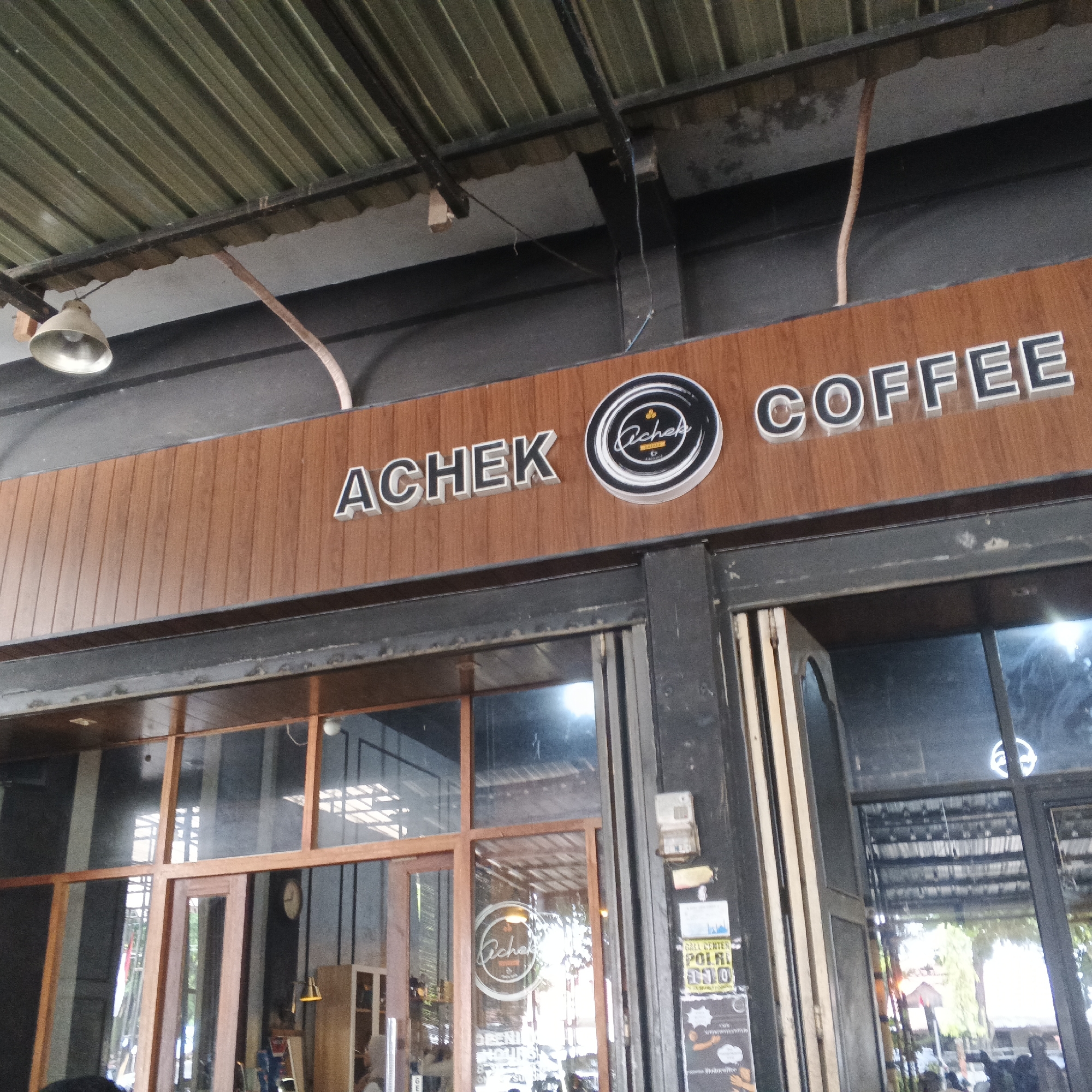The image shows the exterior of a coffee shop named "Acheh Coffee" located in Lhokseumawe, Indonesia. The shop has a prominent wooden sign with the name of the establishment in large, white, 3D letters. A circular logo with the word "Achek" in script is also visible on the sign. The coffee shop appears to have large glass windows and doors, with reflections of the environment visible on the glass. Inside, the interior suggests a seating area. The building has a corrugated metal roof supported by metal beams, and an industrial-style lighting fixture is attached to the underside of the roof. The overall aesthetic suggests a modern yet somewhat rustic design for the coffee shop.