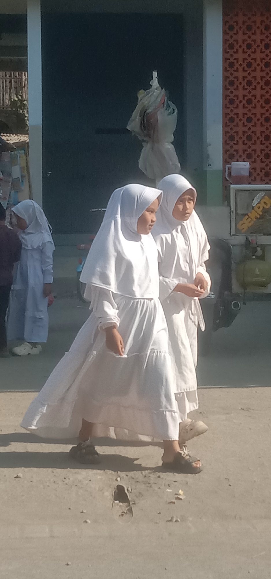 Two young girls, likely students, are walking outdoors in Sukorejo, Indonesia. They are both wearing white Islamic attire, including hijabs that cover their hair and necks, and long, flowing dresses. The girl in the foreground is looking to her right, while the girl behind her is looking forward. They are walking on a dusty, unpaved ground surface. In the background, there is a building with a patterned wall and a dark section that could be a window or a door. To the right of the building, there is a large green cylinder, possibly a gas tank, and some signage with text that is partially visible, including the word "SEMPO." The overall impression is of a casual outdoor scene in a residential or educational setting.