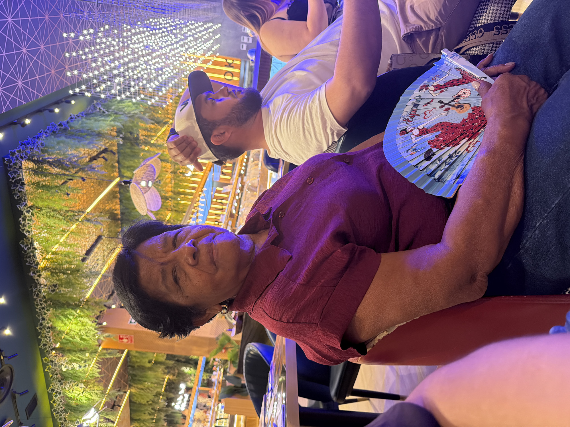 The image shows three people in what appears to be a restaurant or entertainment venue at night. In the foreground, an older woman with dark hair is looking towards the camera, wearing a dark red shirt. To her right, a person's arm is visible, holding a blue fan decorated with images of flamenco dancers. Behind the woman, a man with a beard and a white baseball cap is visible, looking upwards. In the background, there are decorative lights and greenery, suggesting an indoor or outdoor space with elaborate lighting. The overall atmosphere seems relaxed and social. No visible text is discernible in the image.