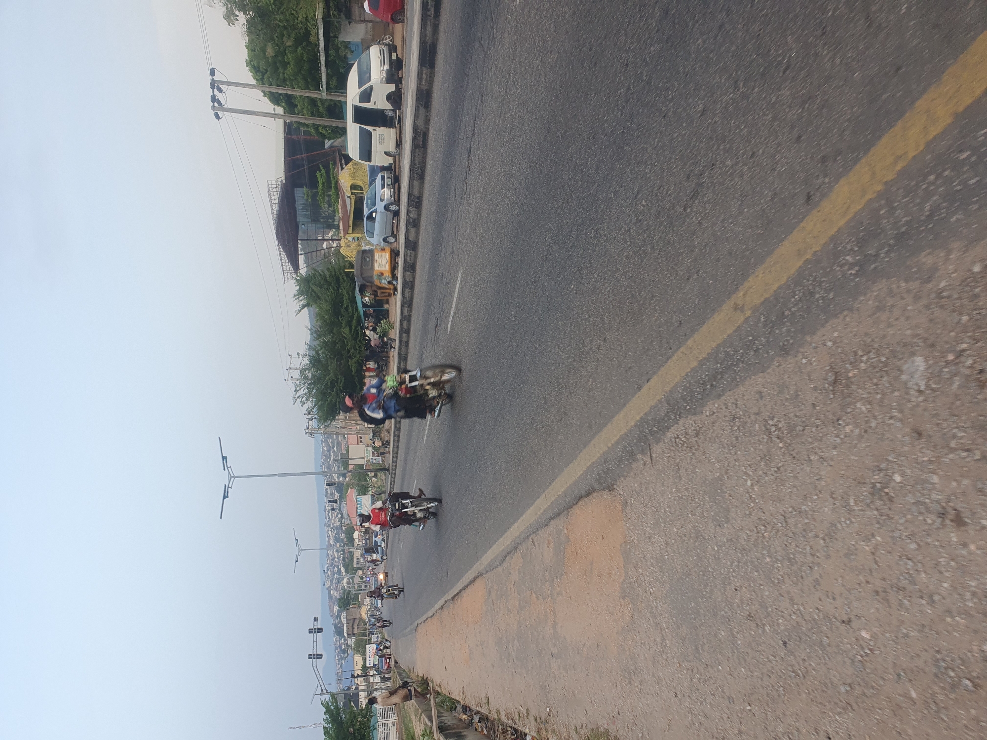 The image shows a street scene in Lokoja, Nigeria, with multiple motorcycles traveling along a paved road. A solid yellow line marks the center of the road. Parked vehicles are visible on the side of the road. Utility poles and street lights are present. The background reveals a cityscape with buildings and vegetation, under a hazy sky.
