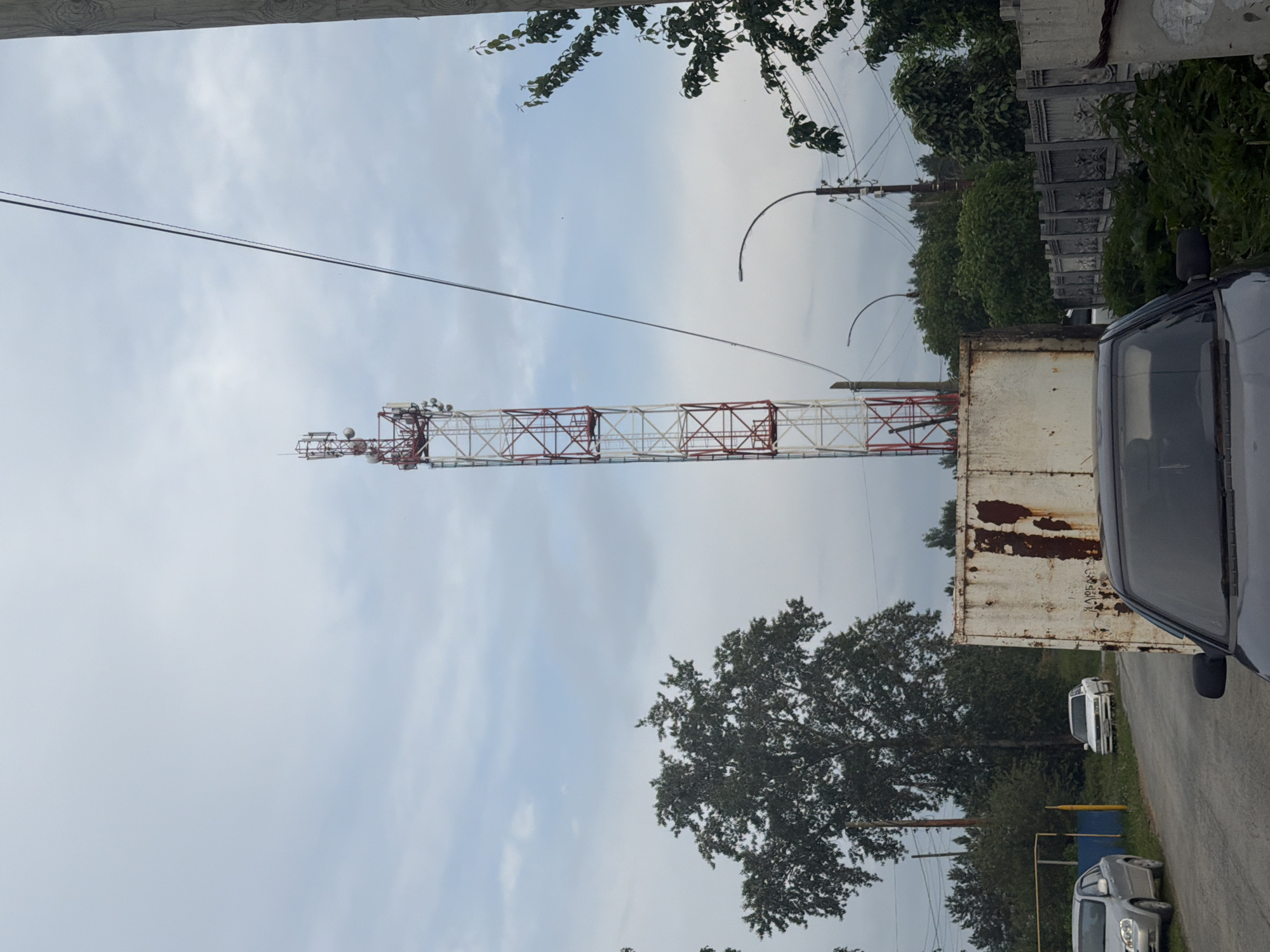A cell tower stands tall in the background, its white and red lattice structure reaching towards a cloudy sky. In the foreground, a rusted metal container sits on the left side of the frame. A dark-colored car is parked in front of it, its windshield visible. To the left of the container, a grey sedan is parked on a dirt road, and a white sedan is parked further down the road. Trees and vegetation line the sides of the road, and a grey decorative fence can be seen in the middle ground on the right. A street lamp arches over the road in the distance.