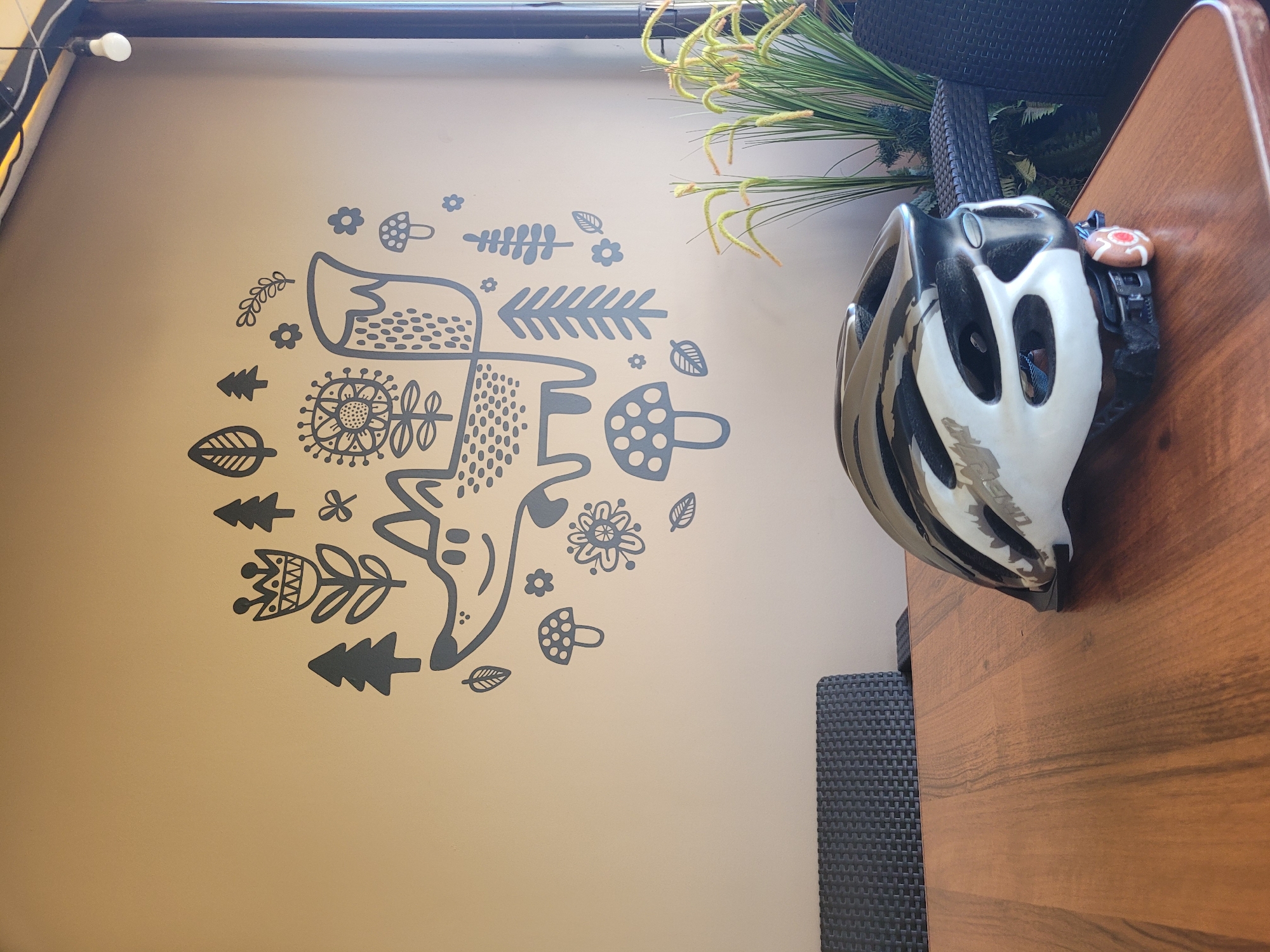 A bicycle helmet rests on a wooden table in what appears to be an indoor seating area. The helmet is white and black with ventilation holes. In the background, a wall is decorated with a large, dark grey graphic depicting a fox surrounded by various stylized natural elements such as trees, flowers, and mushrooms. To the right of the wall decoration, a potted plant with long, grass-like fronds is visible. In the far background, dark-colored outdoor furniture, possibly chairs, can be seen. The image does not contain information that allows for a specific location within the city of Lisy Nos, Russia, to be identified.