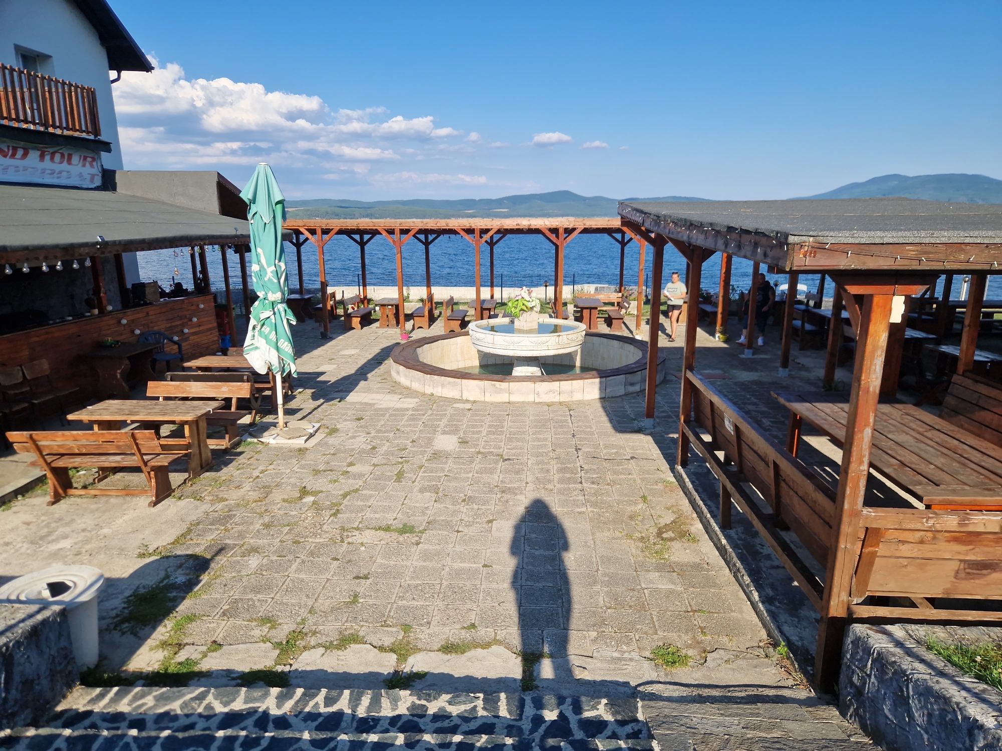 The image depicts an outdoor seating area next to a body of water, likely a lake, on a sunny day with a clear blue sky and scattered clouds. The location is identified as Dolni Pasarel, Bulgaria. The scene features several wooden picnic tables with benches and a circular fountain in the center of the paved courtyard. A green umbrella is propped up near one of the tables.
On the left side of the image, a white building with wooden balconies is visible, with a sign that partially reads "ND TOUR". Adjacent to the building is a covered area with wooden supports, housing more tables and chairs, and decorative string lights. To the right, another covered seating area with wooden structures and roofing is present, occupied by two individuals. One person, wearing a white t-shirt and dark shorts, is walking. Another person, in a light-colored t-shirt and dark pants, is standing.
The ground is paved with square tiles, with patches of grass growing between them. In the foreground, a shadow of a person taking the photo is cast on the pavement. A white cylindrical trash can is also visible near the bottom left. In the background, the calm blue water stretches out to meet rolling, tree-covered hills under a vast sky. The overall impression is of a tranquil, waterside establishment, possibly a restaurant or guesthouse, designed for outdoor relaxation and dining.