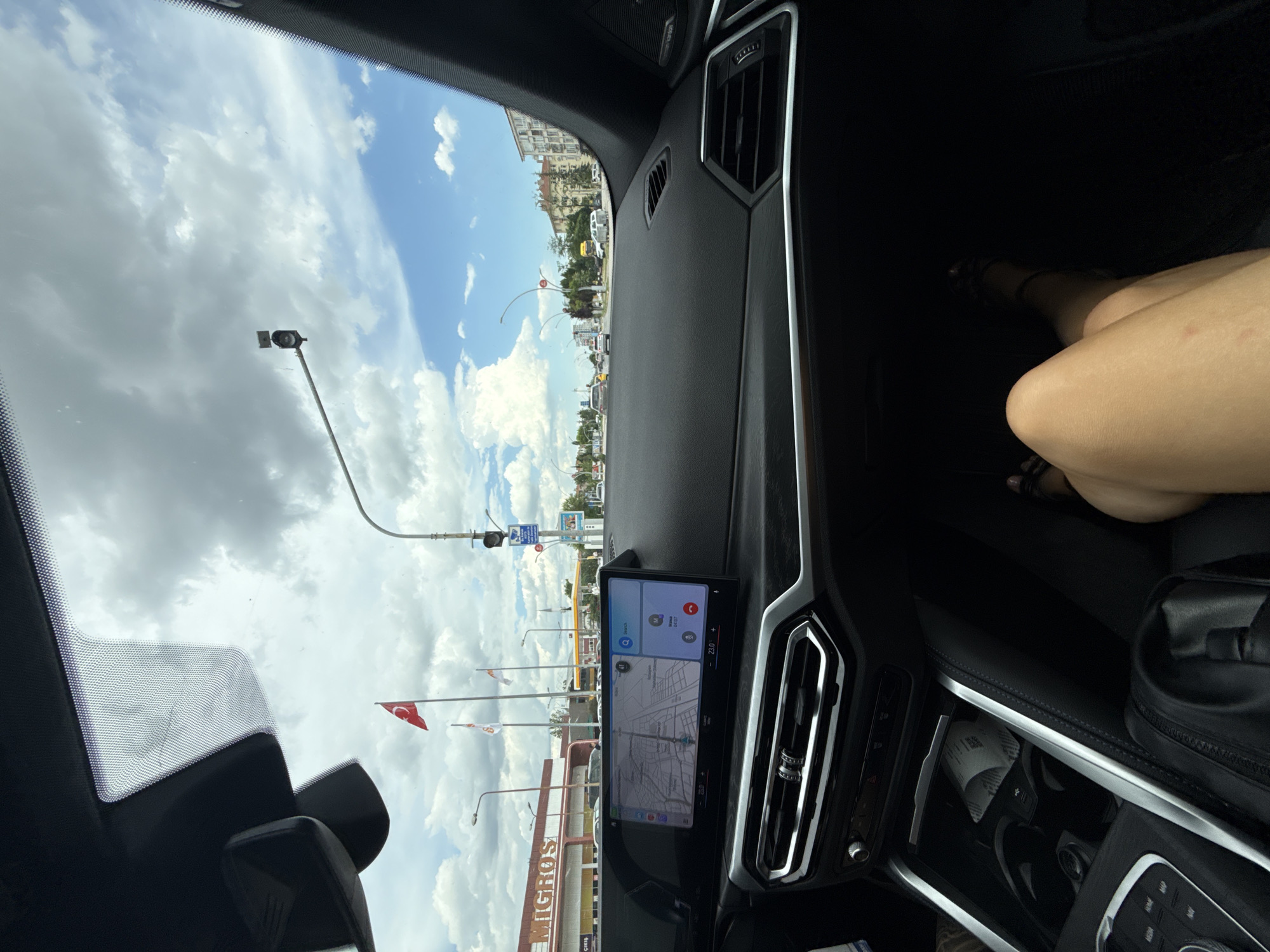 The image, taken from inside a car, shows a view of Edirne Merkez, Turkey. Through the windshield, the sky is visible with a mix of blue and cloudy areas. A traffic light, street lights, and Turkish flags are seen along the road. A building with the name "MIGROS" is also visible. The car's dashboard features a navigation screen displaying a map and controls. Part of the passenger seat is visible, showing a person's leg and a black bag.