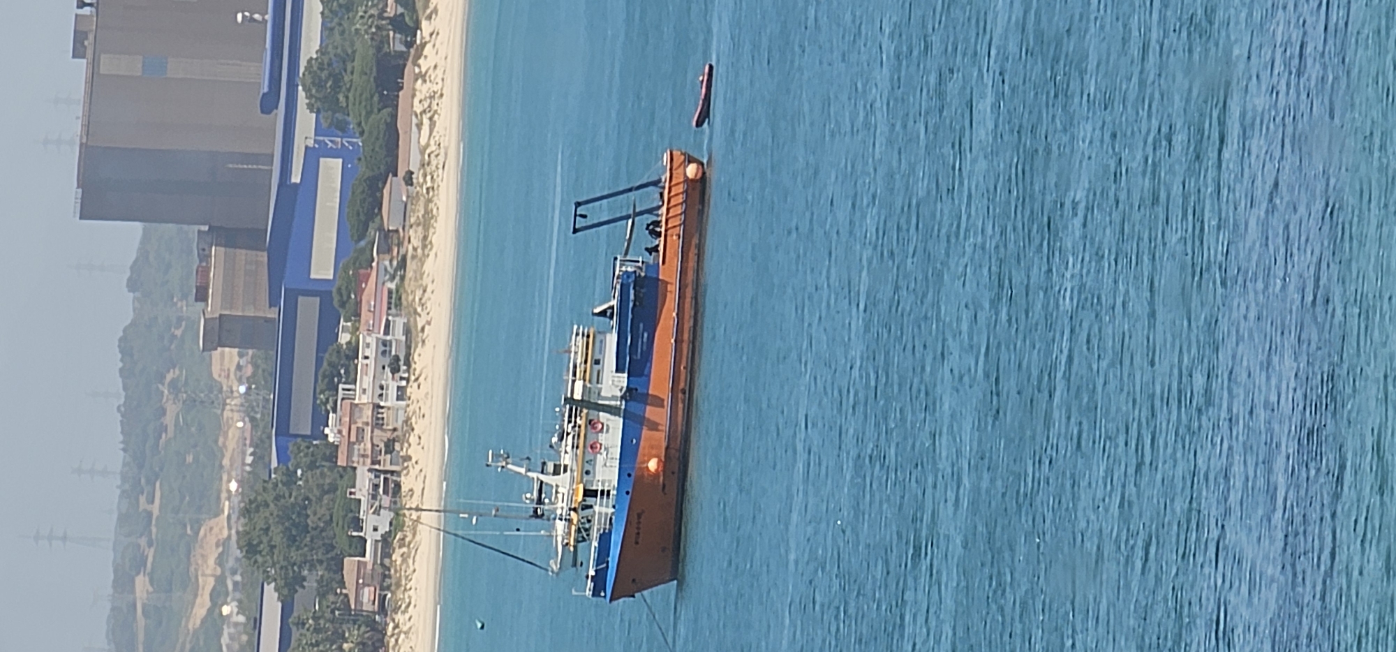 An aerial or elevated view captures a maritime scene in Algeciras, Spain, on a clear day. In the foreground, a large vessel, possibly a fishing trawler or cargo ship, is moored or anchored in calm, blue-green water. The ship features an orange hull and a blue and white superstructure. On the blue section of the hull, the ship's name "DANI-FILA" is visibly inscribed in white letters, with "ALGECIRAS" and some indistinct numbers (possibly "159" or "1594") below it, indicating its home port. A small, red inflatable dinghy or boat floats in the water near the stern of the larger ship.
The sandy coastline extends behind the ship, lined with various buildings, including what appear to be residential houses and possibly small commercial structures, interspersed with green trees and vegetation. Further inland, large industrial buildings or warehouses, some with prominent blue sections, are visible. In the distant background, rolling hills covered in greenery are present, with several power lines or pylons subtly discernible against the hazy sky. The overall impression is one of a bustling coastal area with both industrial and natural elements.