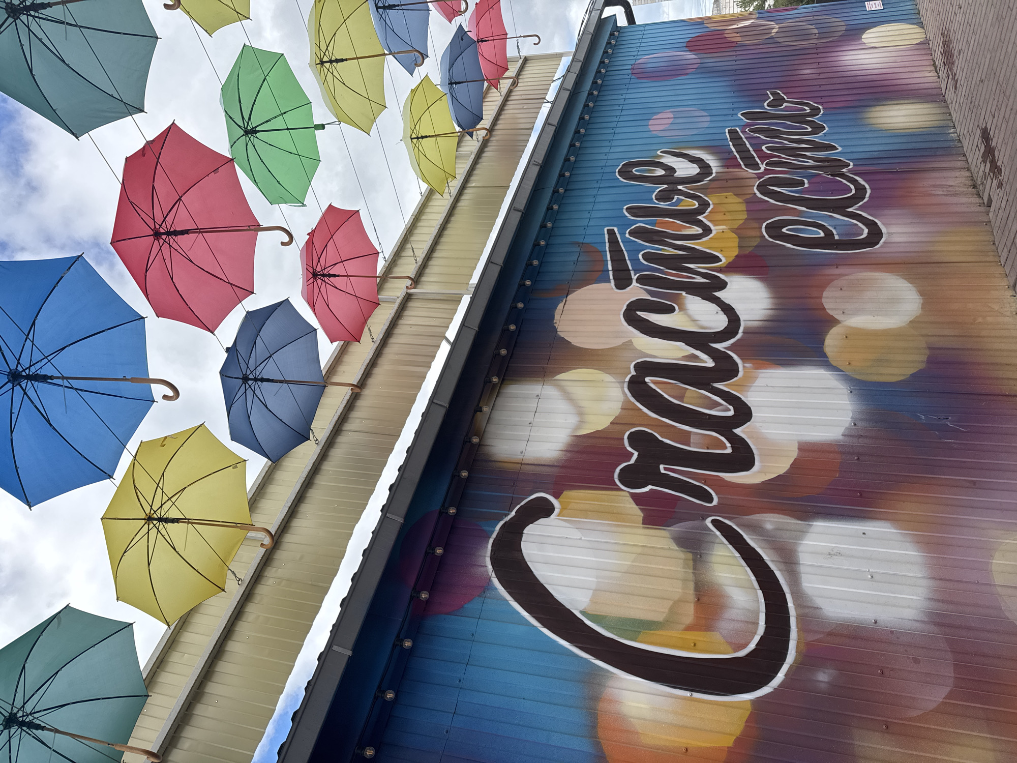 The image depicts an outdoor setting in Osa, Russia, showcasing a decorative display. Overhead, numerous colorful umbrellas of blue, green, yellow, and red hues are suspended, creating a vibrant canopy effect. Adjacent to this is a building wall decorated with a mural that features the Russian phrase "Счастье есть" ("Happiness exists") in stylized black script. The wall surface has a bokeh effect, consisting of blurred circles of color. The overall impression is one of an art installation in a public space.