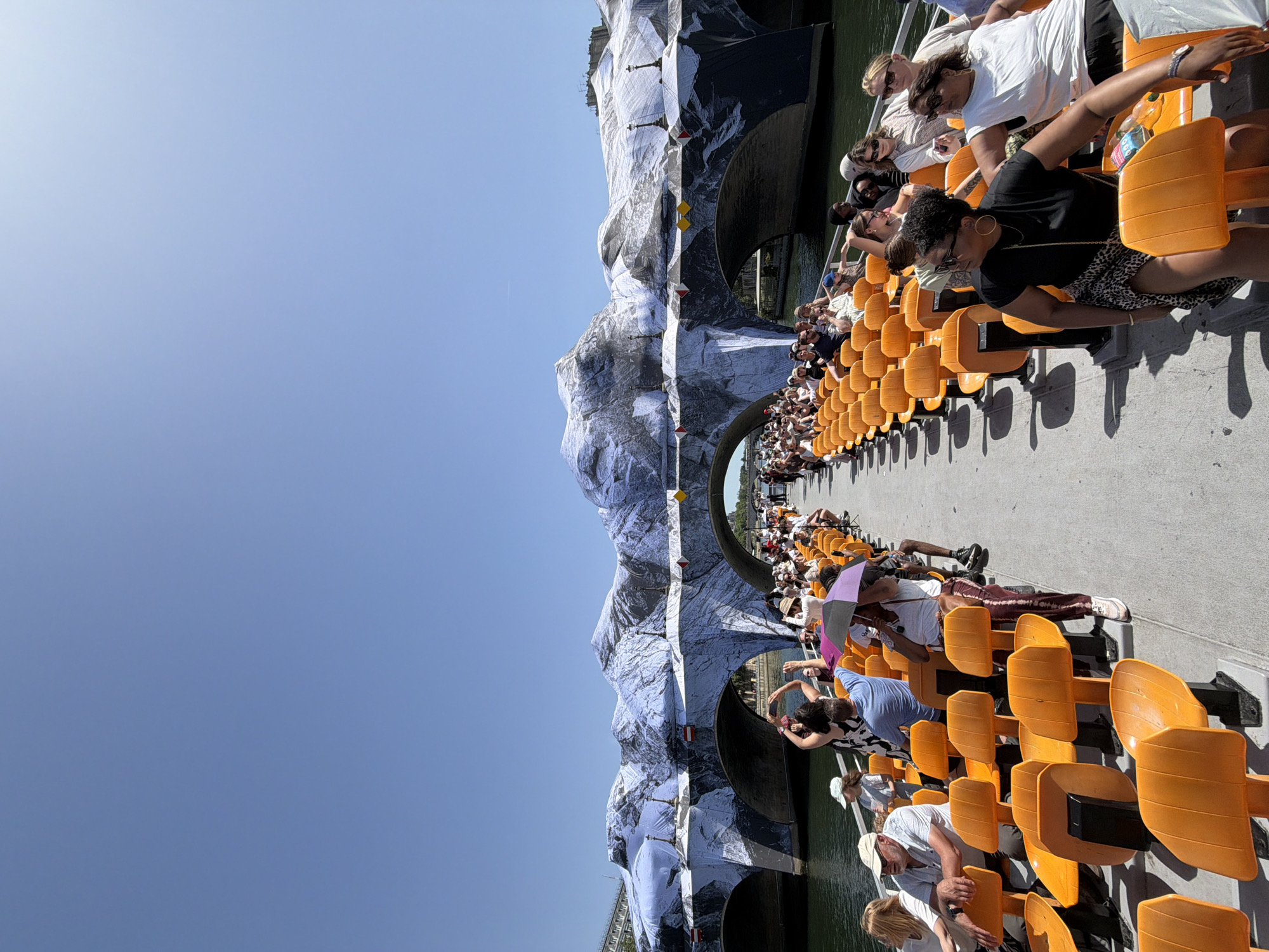 The image shows a long row of orange stadium-style seating along a riverside. Many people are seated, facing towards the right of the frame, with some taking photos. In the background, structures, potentially bridges, are draped in white material printed to resemble mountains. The location is Paris, France, due to the architectural style and river setting, though precise landmarks are obscured by the mountain-like coverings.