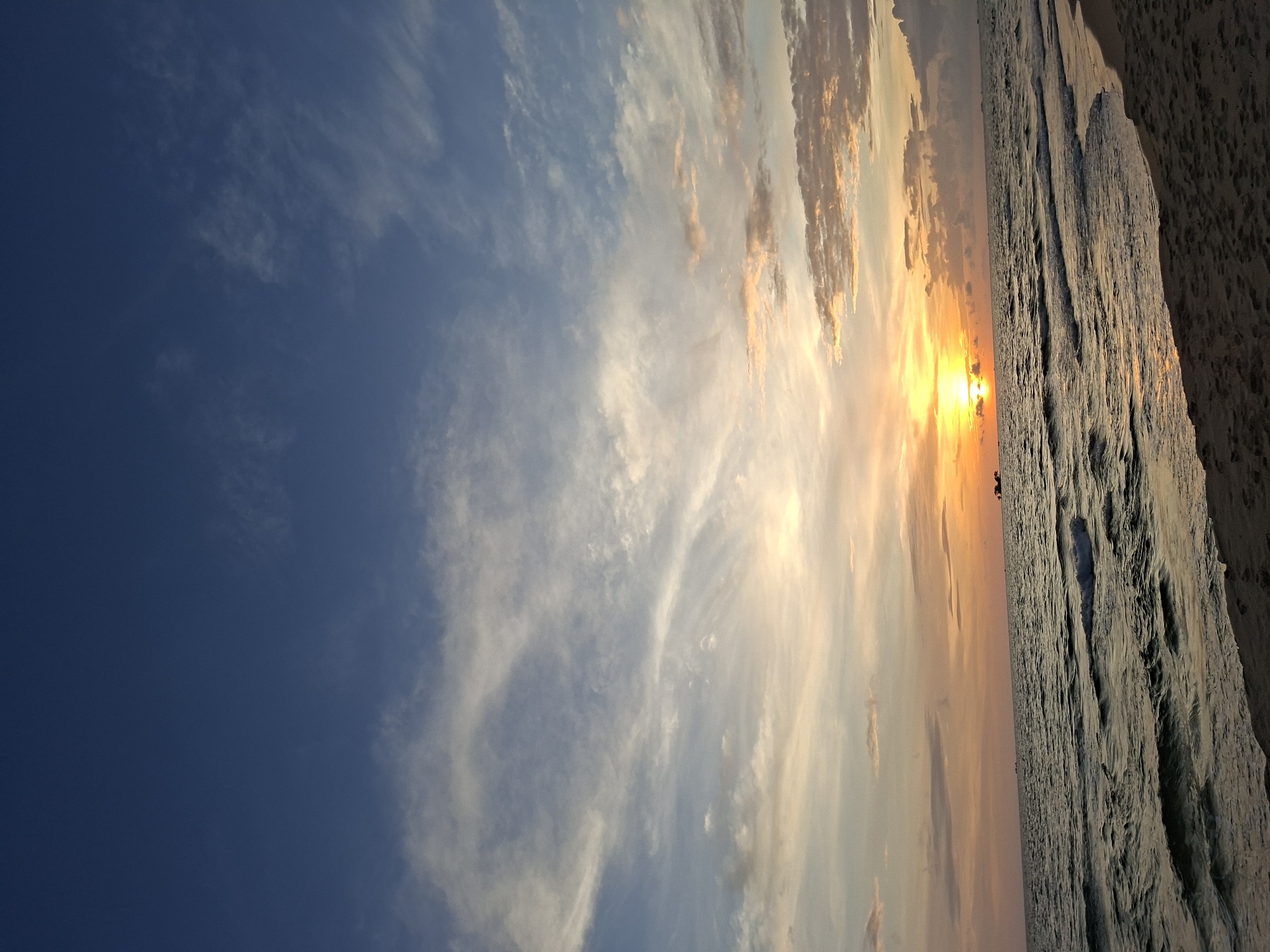 A sunset is captured at eye-level from a sandy beach looking out at the ocean. The sky is a deep blue at the top, transitioning to lighter blues and oranges as it meets the horizon. Thin, wispy clouds are scattered across the sky, some catching the golden light of the setting sun. The sun itself is a bright orb just above the horizon, casting a golden glow on the water. The ocean is dark with small waves lapping at the shore. The sand in the foreground is light brown and textured, showing footprints from previous visitors. In the distance, several small boats are visible on the water. The scene evokes a sense of peace and tranquility.