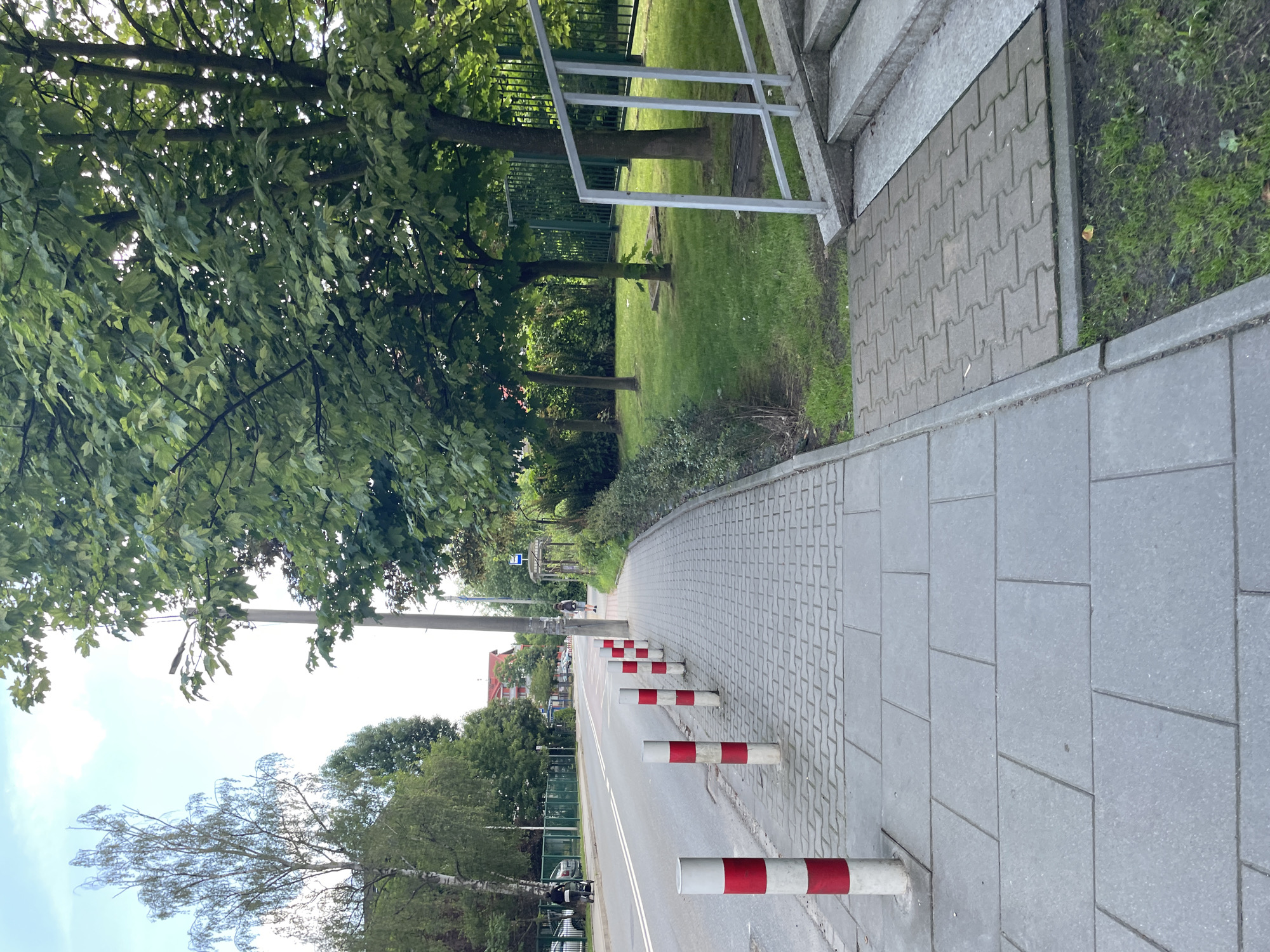 The image depicts a sidewalk in a city, with bollards lining the edge. The sidewalk is paved with gray rectangular tiles, and a section of the path is made of interlocking pavers. Red and white striped bollards are spaced evenly along the sidewalk, separating it from the street. A paved road runs alongside the sidewalk, with a white line marking the lane division. Trees with lush green foliage line the right side of the sidewalk, creating a shaded area. Beyond the trees, a metal fence and grass are visible. In the background, buildings with red roofs and a bus stop shelter can be seen. The sky is overcast with patches of blue visible. There are no people visible in the immediate foreground, but a person can be seen walking in the distance. The overall scene suggests a pleasant urban environment on a cloudy day.