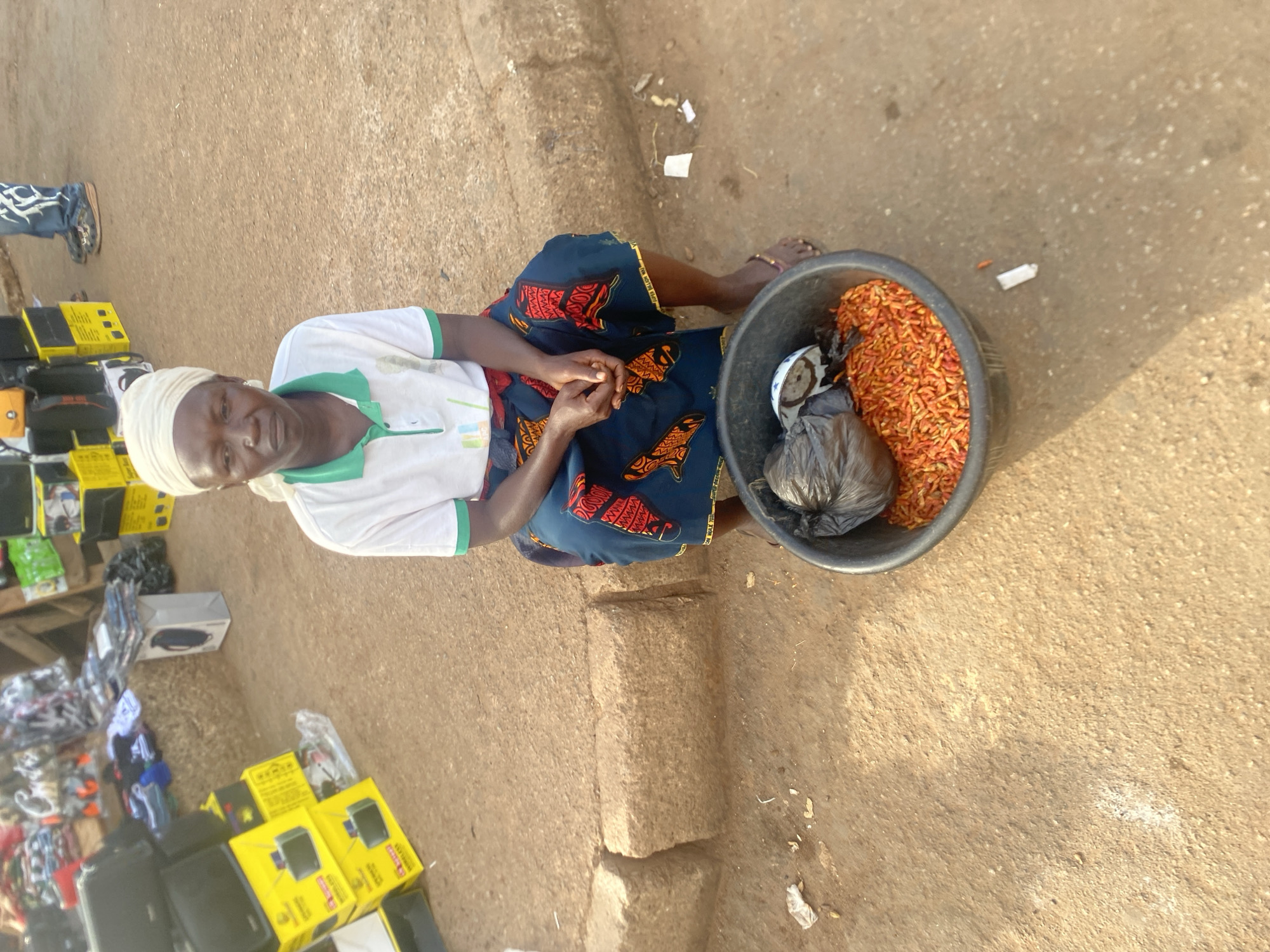 An outdoor, eye-level shot captures an elderly woman sitting on the ground, likely in a market or roadside stall in Bukuru, Nigeria. She wears a white headwrap, a white polo shirt with a green collar, and a blue patterned wrapper skirt. Her hands are clasped in front of her, and she looks calmly at the viewer.
Beside her, to her right, is a large black bucket brimming with bright red-orange chili peppers. Inside the bucket, a small white bowl and a crumpled black plastic bag are also visible. Behind and to her left, a makeshift stall displays various goods. Numerous yellow boxes, possibly containing electronic accessories like chargers or power banks, are stacked. Text such as "WIRELESS" is faintly discernible on these boxes, along with other unreadable text. Other items, including black speaker-like devices and plastic-wrapped products, are also on display. Part of another person's leg and foot, wearing blue jeans and shoes, can be seen behind the stall.
The ground is unpaved, consisting of reddish-brown dirt, with some scattered litter. Bright, direct sunlight casts clear shadows, indicating it is daytime, likely midday or early afternoon, with clear weather. The scene portrays a common market setting with a vendor selling produce and other goods.