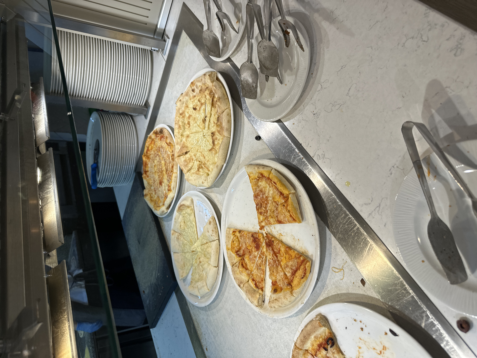 The image shows a self-service buffet, likely in a restaurant or hotel. The buffet line features several plates of sliced pizza. There are several types of pizza, including cheese and pepperoni. Next to the pizzas are stacks of white plates. Utensils, including tongs and serving spoons, are also present for customers to serve themselves. The environment is clean and well-lit. The location is unspecified in the image. There is no evidence of people or specific actions. The image's lack of context makes determining a specific location such as Kazanlak, Bulgaria impossible.