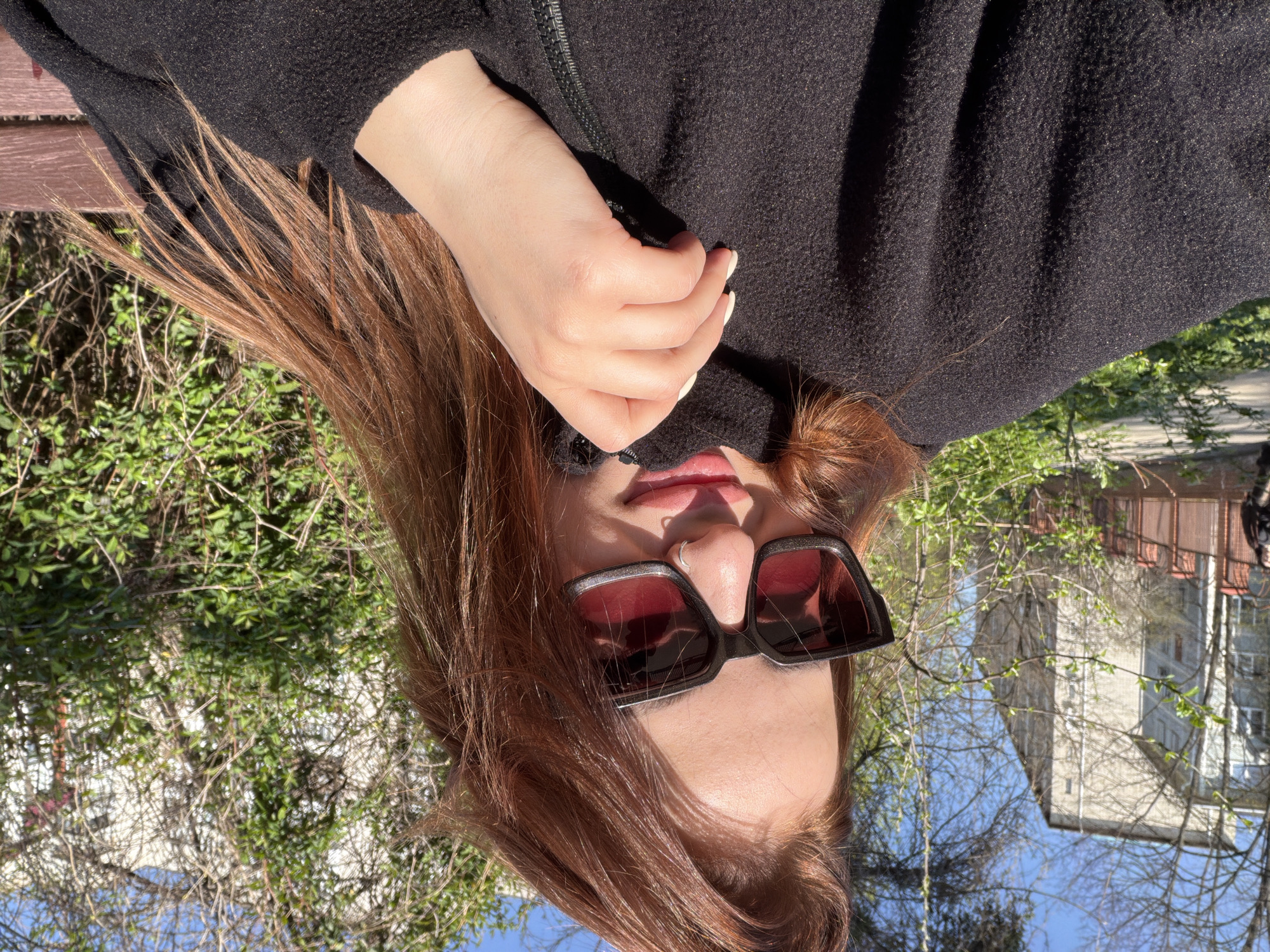 A woman wearing black sunglasses and a black jacket sits on a wooden bench outdoors. The image is taken from an unusual angle, appearing upside-down. The woman has long, auburn hair. Her right hand is visible near her chin. In the background, vegetation and buildings are visible. Based on available visual evidence, it's not possible to confirm the exact location in Krasnodar, Russia.