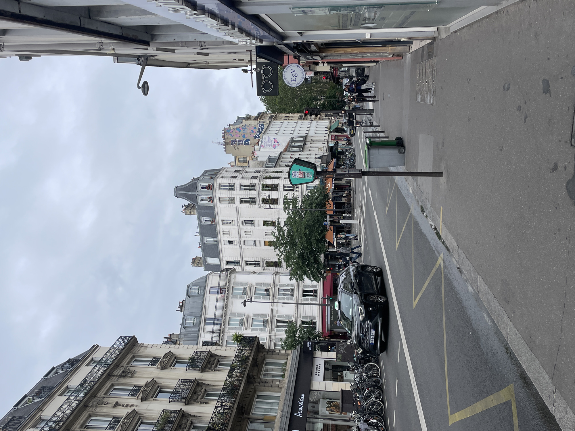 This street-level photograph captures a typical urban scene in Paris, France, under an overcast sky during daytime. The image, rotated vertically, showcases a bustling street flanked by classic Haussmannian buildings with ornate balconies, detailed facades, and mansard roofs featuring dormer windows.
On the left side, the buildings display intricate wrought-iron railings. Towards the center and right, more buildings are visible, including one with extensive colorful graffiti, prominently featuring the word "BUGS" alongside abstract designs. Shopfronts line the ground floors of these structures. Visible signage includes "Acousticien," "ISABEL MARANT," "AFFLELOU," and "Toilettes H." A black circular sign with "B&V" and "BOULANGERIE ARTISANALE" indicates a bakery, while a square black sign with "E&V" is also seen.
The street itself is asphalt with yellow zigzag markings indicating a no-parking zone, along with white lines. A black SUV with the French license plate "GB-846-HY" is parked on the street. Alongside the curb, numerous bicycles are neatly parked in a bike rack, and several scooters or motorcycles are also visible. Green public bicycle rental stands and trash bins are present on the sidewalk. Traffic lights, currently red, and streetlights are part of the urban infrastructure.
Several pedestrians can be seen on the sidewalks, moving about their daily routines, contributing to the everyday life of the city. A security camera is mounted high on a building. The overall atmosphere is one of a busy, yet ordinary, Parisian street scene on a cloudy day.