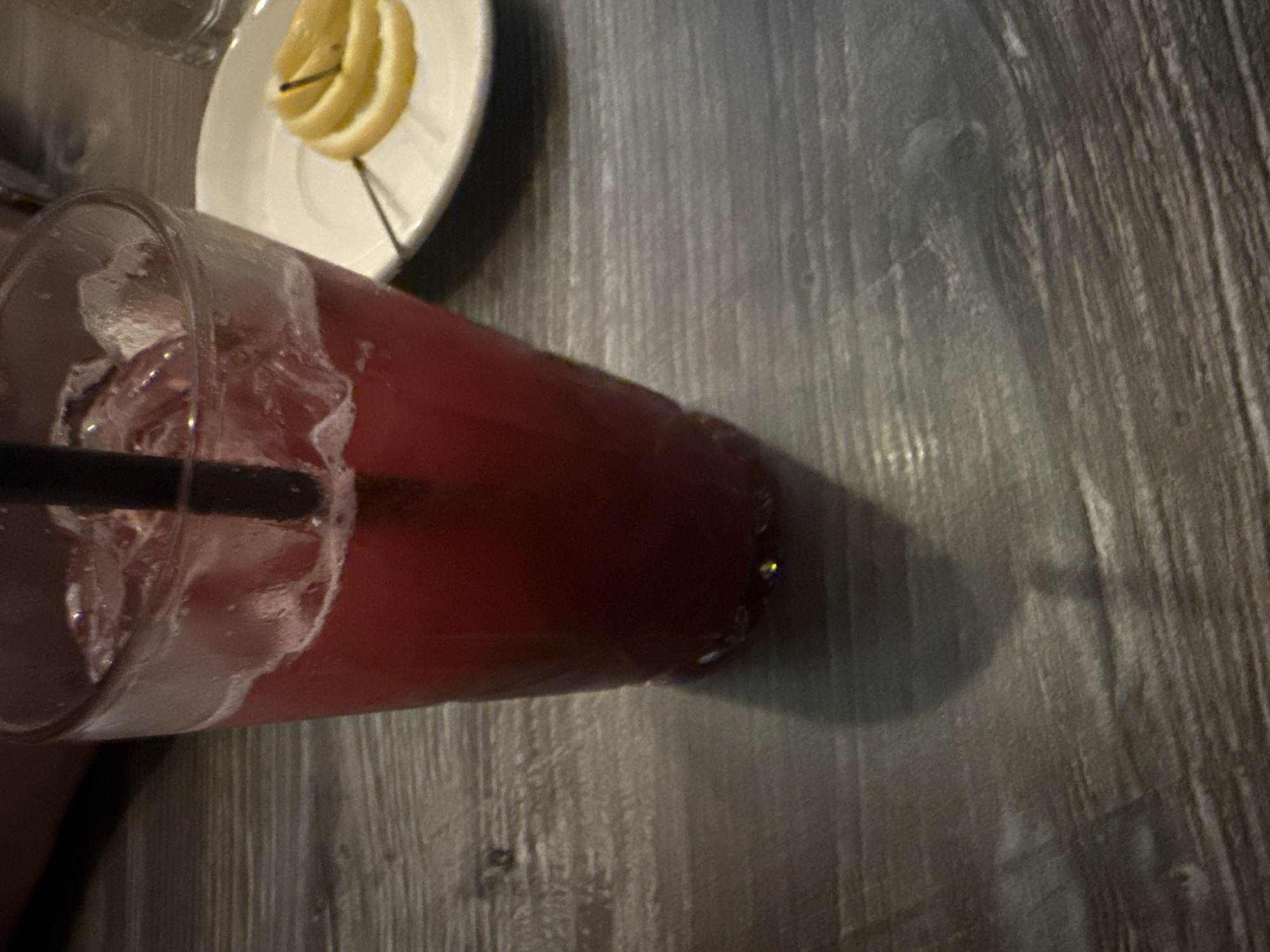 A close-up, overhead shot depicts a glass of red liquid, likely a beverage, filled with ice and a black straw. The glass sits on a gray, textured surface, possibly a wooden table. A small white plate with several lemon slices is partially visible in the upper left corner of the image. The location in Glenview, United States of America, cannot be determined from the image alone.