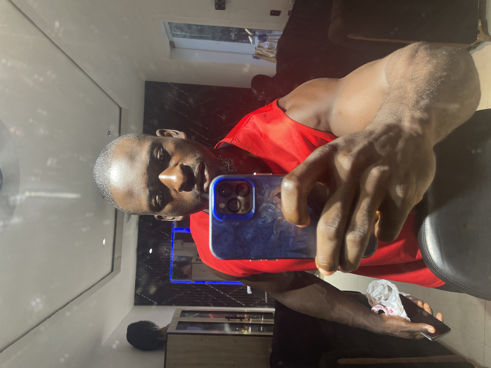 A man is taking a selfie in what appears to be an indoor location. He is wearing a red tank top and holding a smartphone with a blue and patterned case. He is looking at the camera. In the background, there's a cabinet and what might be a television with a blue light emitted from it. The location is unknown, and cannot be determined as Akwanga, Nigeria from the visual data provided.