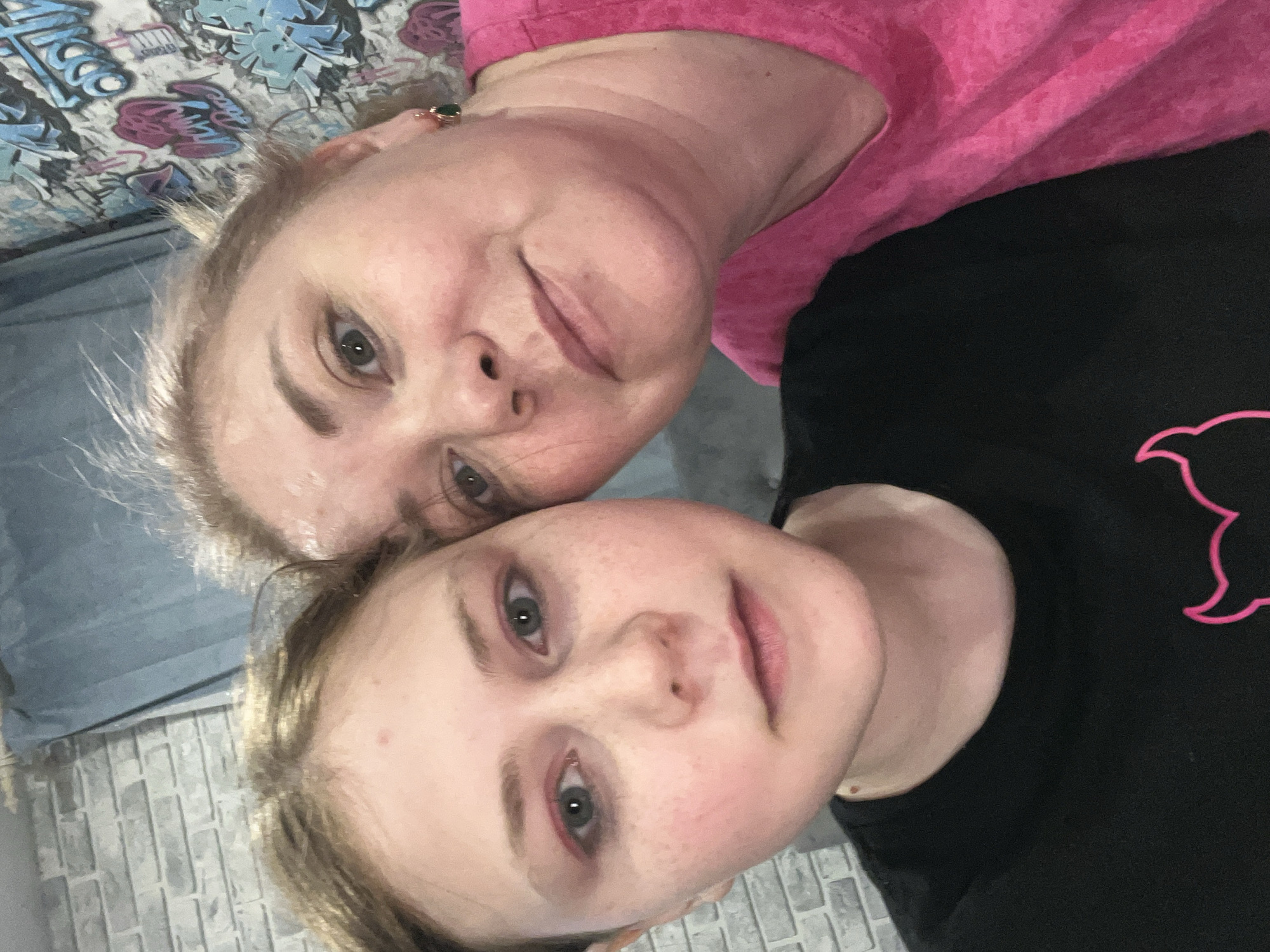 A selfie shows two people: an adult and a child. The adult has blonde hair and is wearing a pink t-shirt. The child has blonde hair and is wearing a black t-shirt with a pink outline design. The background includes a bed with a patterned comforter and a brick-patterned wall. The location is possibly Vesyoloye, Russia. There is no explicit action or interaction.