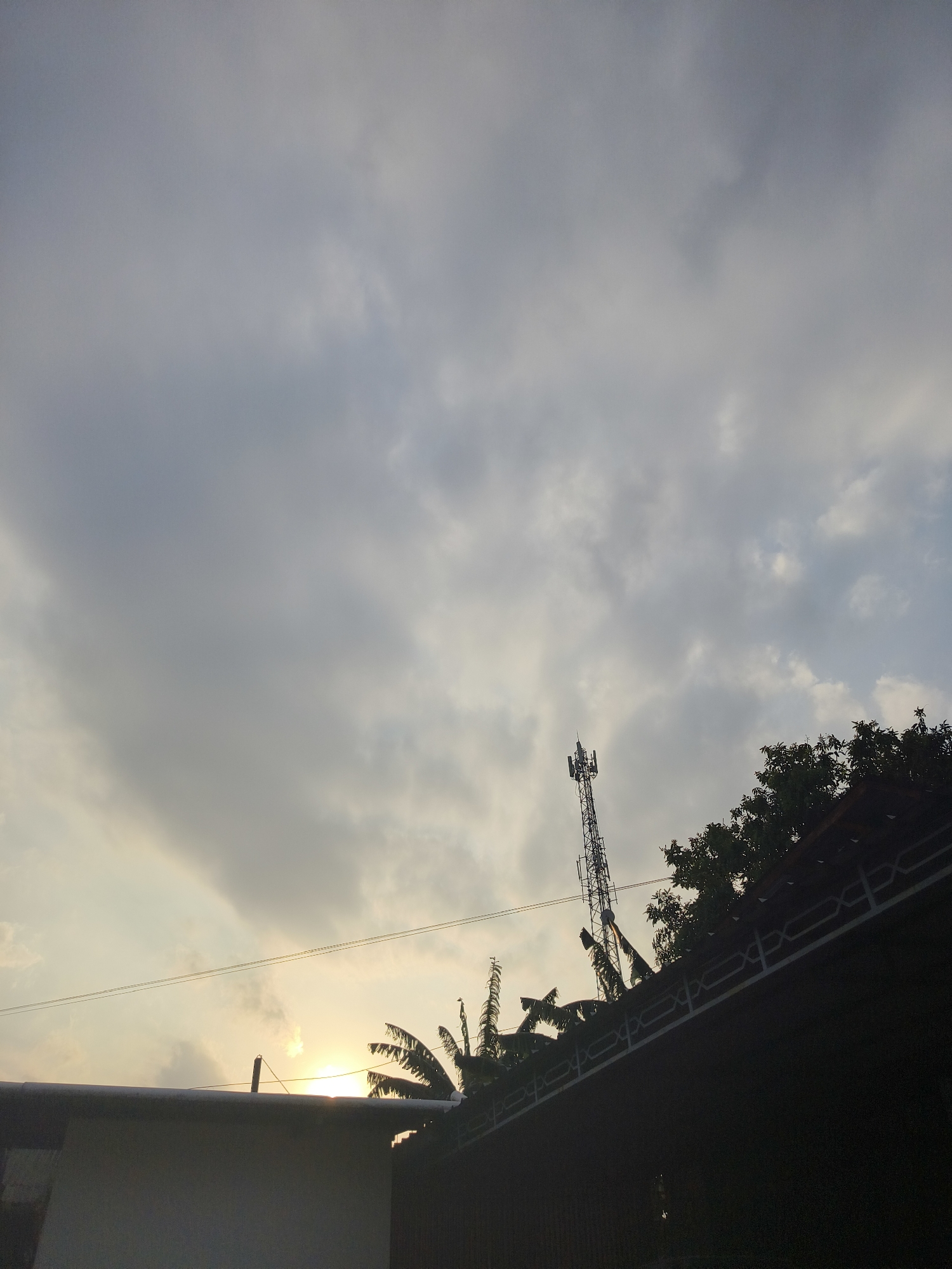 The image depicts a silhouetted view of a cell tower against a cloudy sky at sunset. In the foreground, the dark silhouettes of rooftops, trees, and utility wires are visible. The sun, a bright orb, is partially obscured by clouds and peeking through the leaves of a palm tree on the right side of the image. The overall scene suggests an urban or suburban environment in Pangkalpinang, Indonesia, likely captured during the golden hour.
