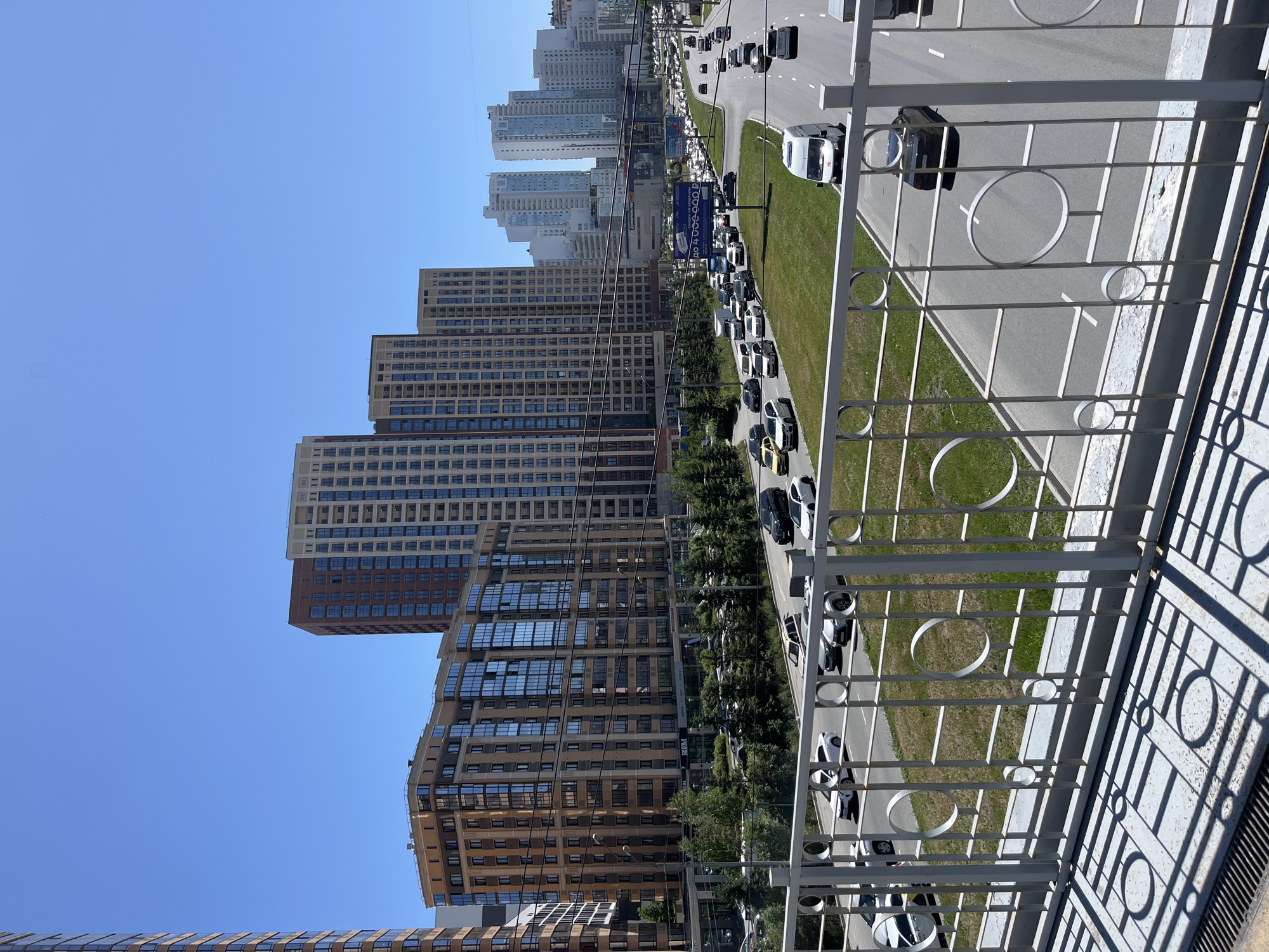The image captures a wide, multi-lane road in an urban setting, likely a city. The road is congested with traffic, with numerous cars and vans visible. Several tall, modern apartment buildings with glass facades dominate the skyline, suggesting a residential area. Green trees and a grassy median separate lanes of traffic, indicating landscaping within the city. The sky is clear and blue, suggesting a sunny day. A white metal railing is in the foreground, casting shadows on the ground, implying the photo was taken from a higher vantage point, possibly a bridge or overpass. There are no people clearly visible in the image. The overall scene depicts a busy urban environment during daylight hours.