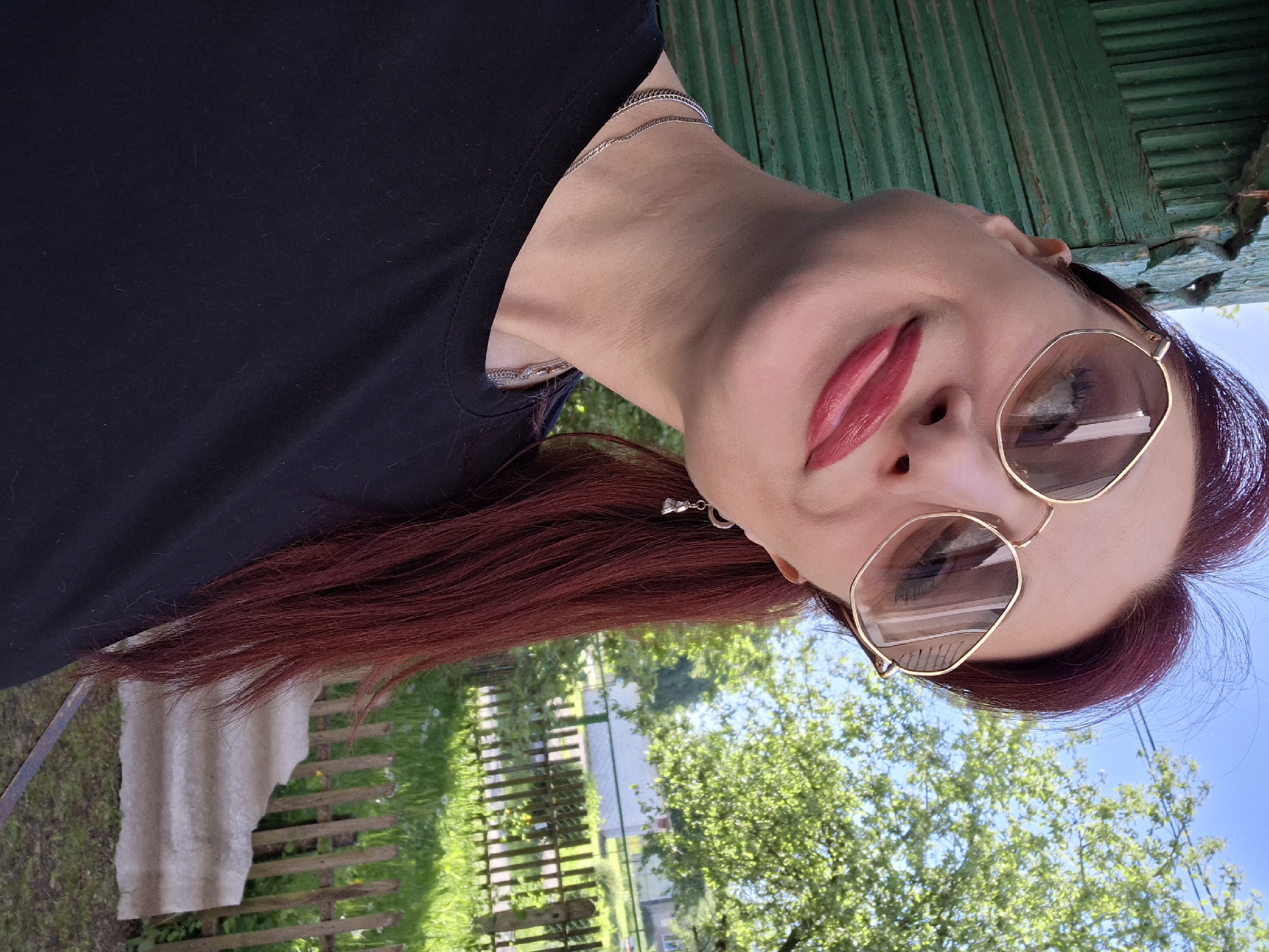 A woman with burgundy hair is facing the camera at an angle. She wears sunglasses with gold frames and pink lipstick. She has a black shirt and a silver necklace. In the background, there is green foliage, a fence, and a green wooden structure. The location is Забалоцкі сельскі Савет, Belarus.
