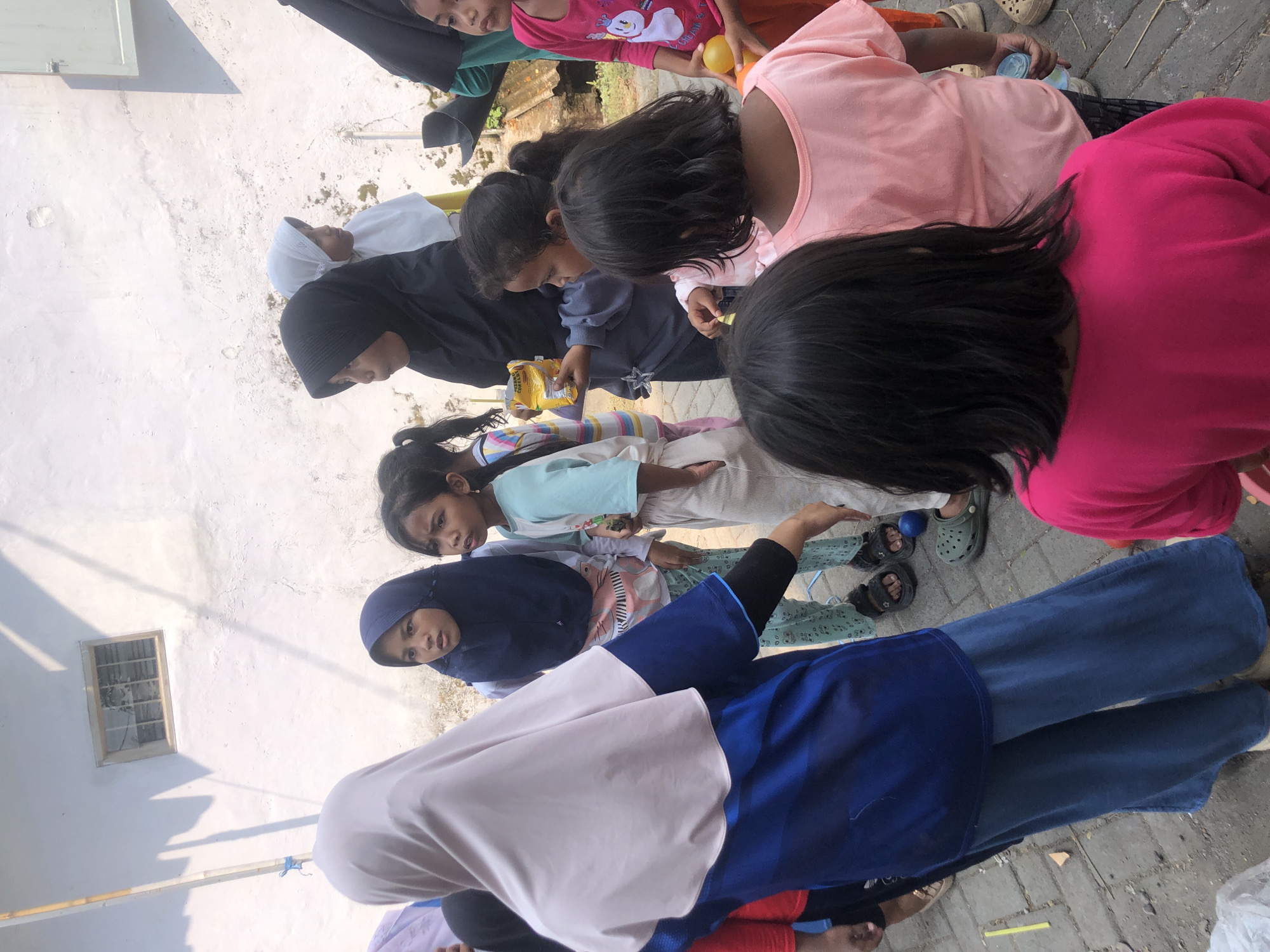 A group of children and adults are gathered outdoors on a paved surface, likely a street or courtyard in Tenggilis Rejo, Indonesia. The scene is illuminated by natural daylight. A wall with a small, barred window forms the backdrop. Several children are the primary subjects, dressed in casual clothing, with some wearing hijabs. One adult, wearing a light pink hijab and a blue and black outfit, stands to the left, facing away from the camera. Another adult, in a black hijab, is visible in the center. The children appear to be engaged in an activity, possibly a game or a distribution of snacks. One child in the center is holding a yellow snack packet. Another child on the right is holding a yellow ball. The ground is paved with rectangular stones and some debris is scattered around. The overall impression is a lively gathering of people in a residential or community area.