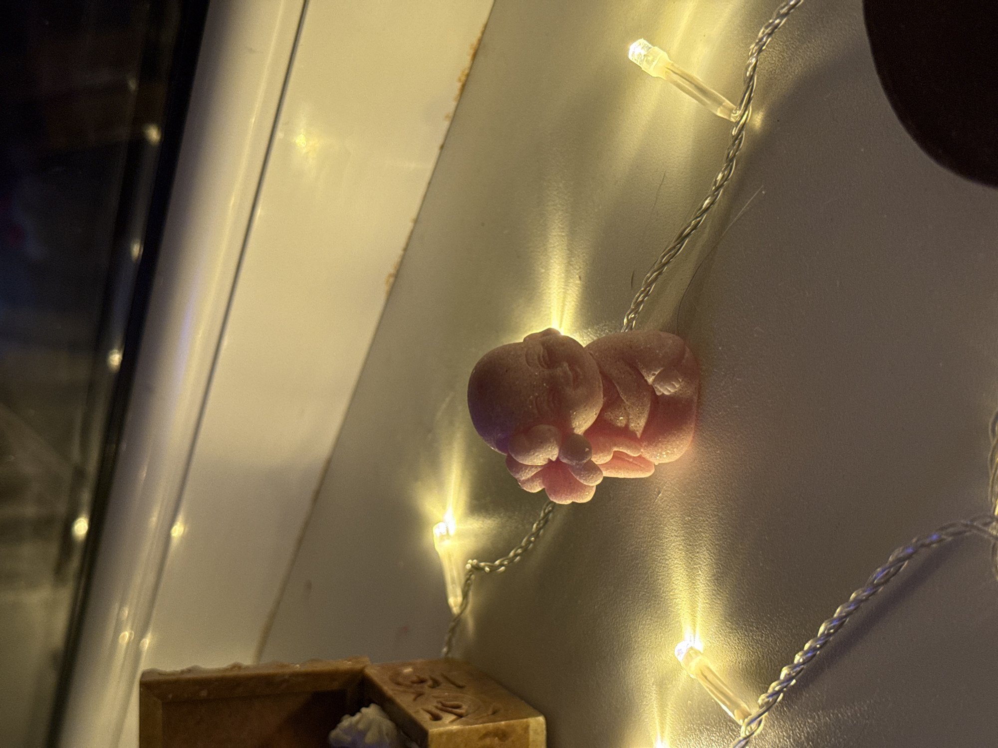 The image shows a close-up of a small, pink, glittery figurine resembling a baby with its hands near its face, sitting on a white surface. A string of warm white LED lights is draped around the figurine, with several lights illuminated and casting a glow. To the left of the figurine is a small, ornate wooden box with a lid that is partially open, revealing white material inside. The background is out of focus, but suggests a window sill or shelf with reflections of lights. There are no people visible in the image. The location of this scene in the city of Naberezhnye Chelny, Russia, is not discernible from the visual information provided in the image.