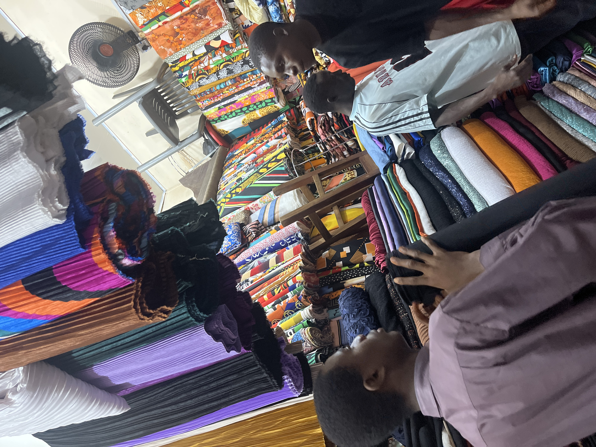 The image depicts the interior of a fabric store, likely in Jos, Nigeria, characterized by abundant rolls and stacks of vibrantly colored textiles. In the foreground, a young Black male is seen from behind, wearing a muted purple collared shirt, his right hand reaching out towards a pile of dark fabric. To his right, another young Black male, facing away from the camera, wears a light blue t-shirt with dark stripes on the sleeves and a red graphic on the front. He is looking towards the right and his right hand is gesturing downwards. In the background, a black oscillating fan is mounted on the wall. The store is filled with a variety of fabrics, including pleated materials in shades of white, purple, brown, green, and multi-colored patterns. Further back, a rack displays numerous rolled fabrics with intricate designs and bold colors, such as orange, yellow, blue, and black patterns. The lighting is dim, suggesting an indoor setting without direct sunlight. The overall impression is of a busy marketplace stall overflowing with a wide selection of fabrics.