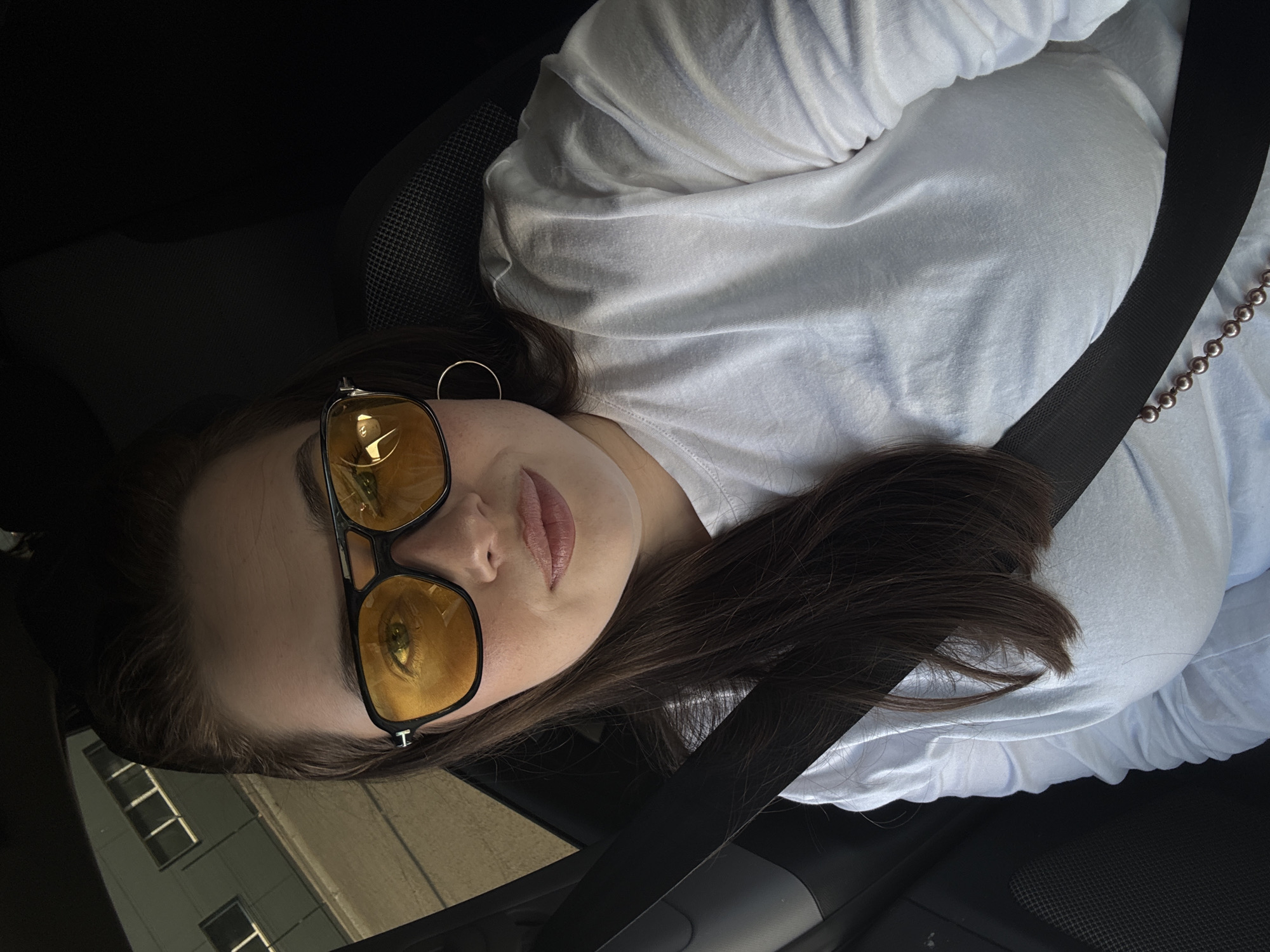 A woman with long, dark hair is wearing yellow sunglasses and a white t-shirt, secured with a black seatbelt. She is inside a car. Reflections in her sunglasses indicate a building with windows. The location appears to be the city of Chaikovsky, Russia.