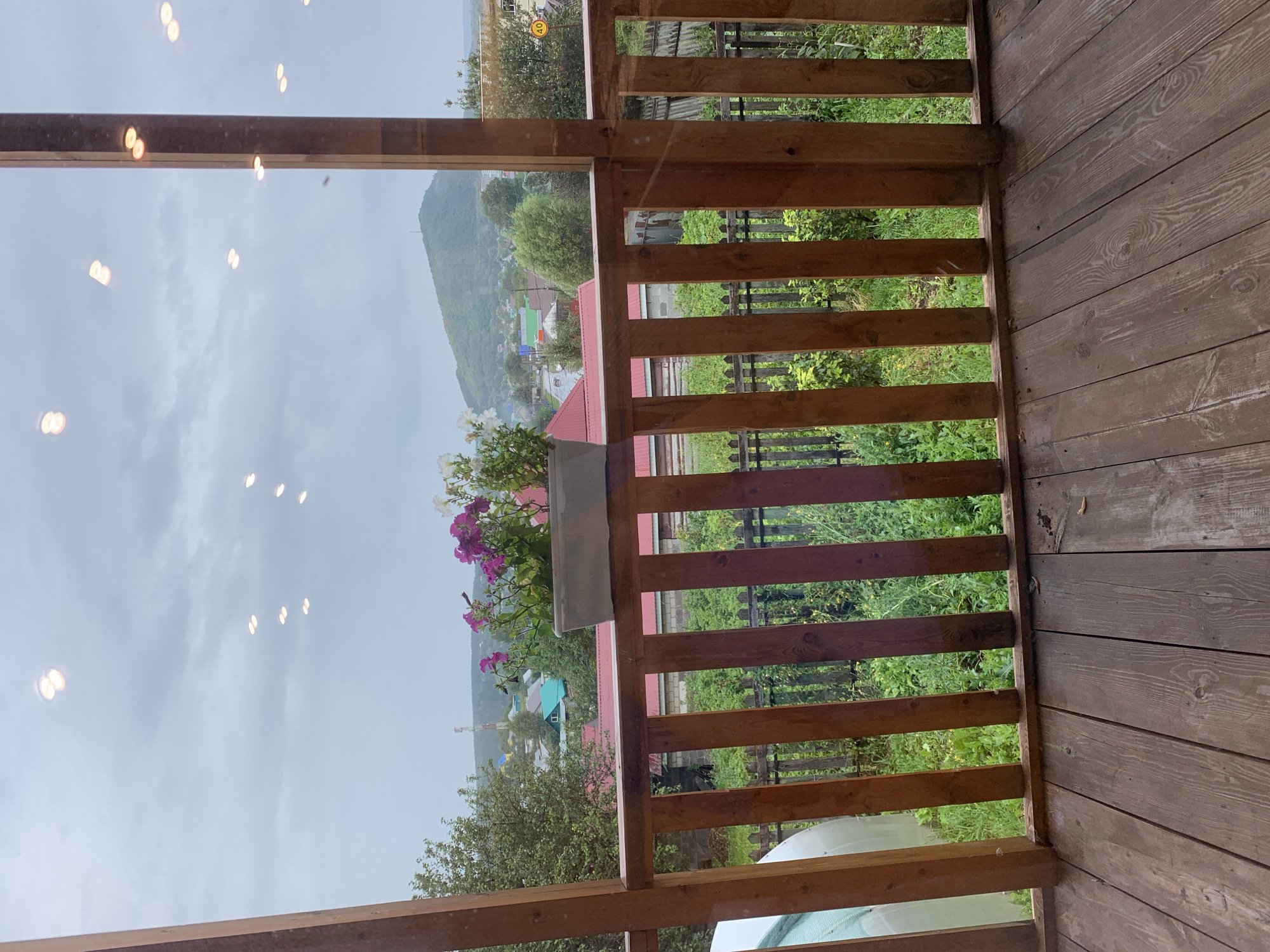 The image shows a view from a wooden porch overlooking a suburban landscape in Asha, Russia. The foreground consists of weathered wooden floorboards and a wooden railing. Beyond the railing, a narrow garden with green foliage is visible, bordered by a rustic wooden fence. In the middle ground, various residential buildings with red and green roofs are scattered amongst trees. A prominent hill with a communication tower on its summit dominates the background. The sky is overcast with scattered, blurred lights, possibly reflections on a window. A planter box with pink and white flowers sits on the railing. The overall scene depicts a quiet, rural residential area under a cloudy sky.
