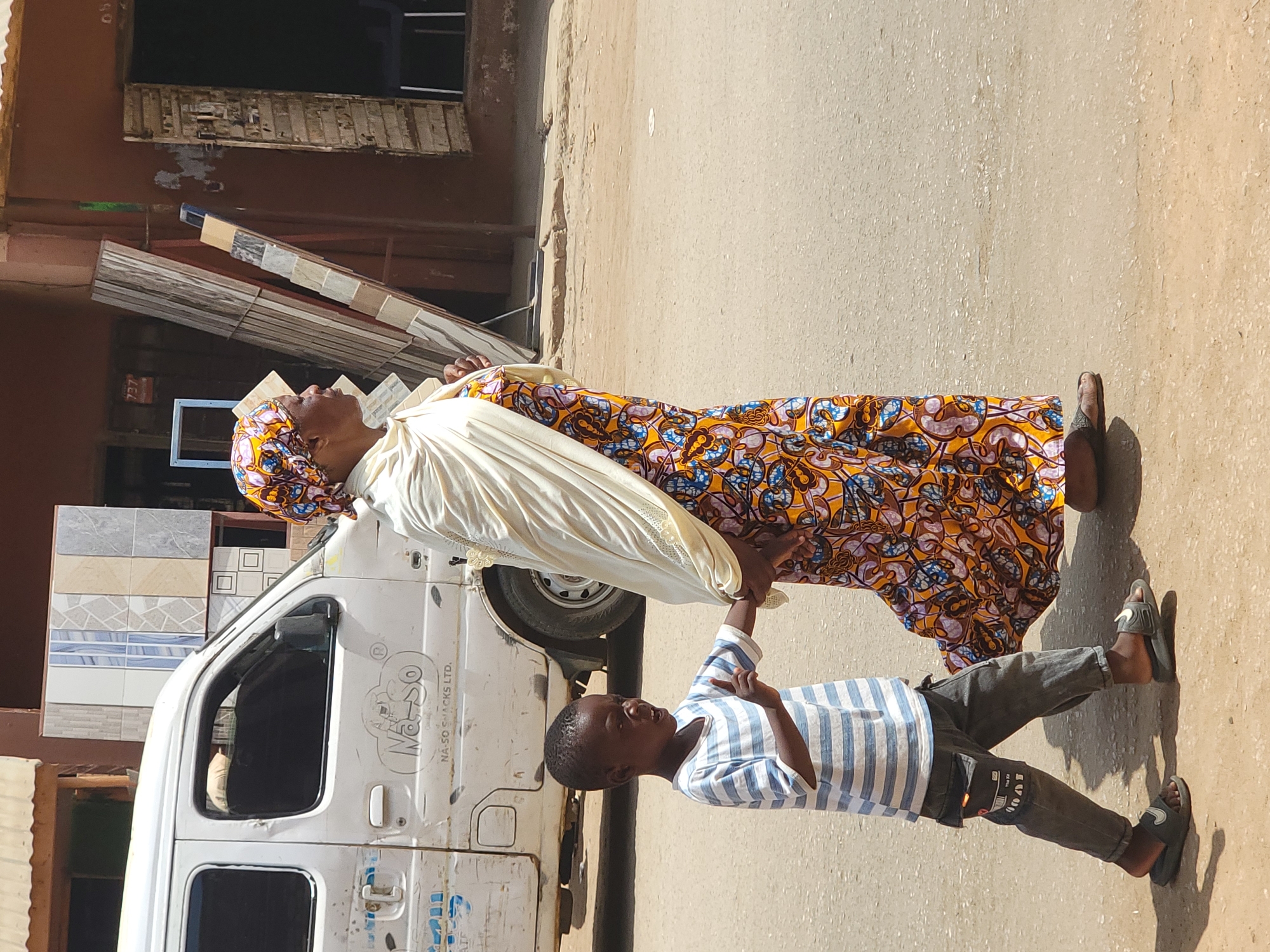 An eye-level outdoor shot depicts an elderly woman and a young boy standing on a paved street in Maiburiji, Nigeria. The woman, wearing a vibrant, patterned orange and blue dress and a matching head wrap, has a white shawl draped over her shoulders. The boy, clad in a blue and white striped shirt and gray pants, holds her hand. They stand adjacent to a white van with tiles stacked on top. The background reveals buildings with windows and various construction materials. The scene is bathed in bright daylight.