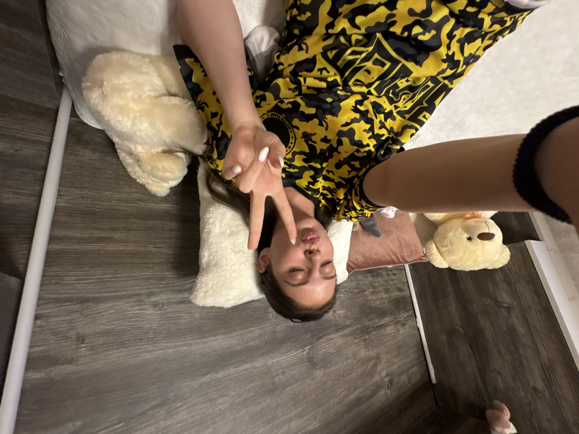 A young woman, appearing to be in her late teens or early twenties, is lying on a bed, holding up her right hand in a peace sign and puckering her lips. She is wearing a yellow and black camouflage patterned t-shirt. The bed is covered with white bedding. Behind her, there are several stuffed animals, including a large light brown teddy bear and a smaller cream-colored teddy bear. The wall behind the bed has a wood-paneled texture. The scene is indoors, possibly a bedroom, in the city of Bulgarovo, Bulgaria.