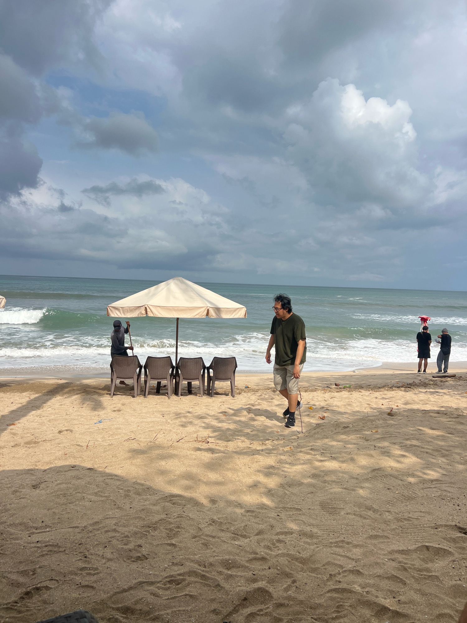 A beach scene in Kuta, Indonesia, features a sandy shore with gentle waves rolling in from the ocean under a cloudy sky. In the foreground, a man wearing a dark green t-shirt and light-colored shorts walks across the sand. Behind him, a beige umbrella is set up, with four empty plastic chairs arranged underneath it. To the left of the umbrella, a person wearing dark clothing and a cap stands with a long pole. Further down the beach, near the water's edge, three individuals are visible: one wearing a black shirt and shorts, another in a black shirt and grey pants, and a third in a red hat and black shirt. The overall atmosphere suggests a moderately busy beach day with overcast conditions.