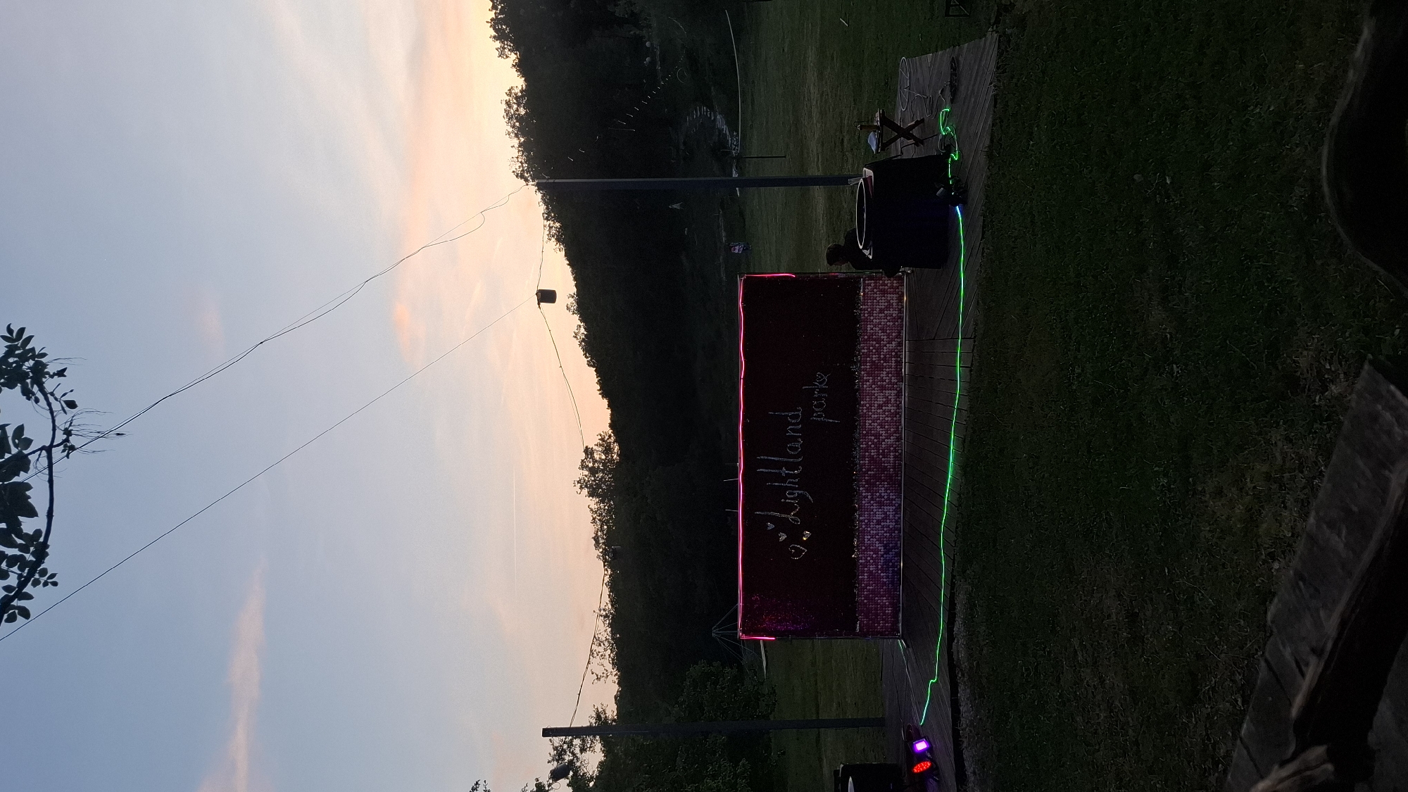 The image shows an outdoor event in Bukovik, Montenegro. A sign reading "J. Lightland parke" is illuminated with pink and green neon lights. The sign is mounted on poles and positioned on a wooden platform. A person is visible sitting on the platform near audio equipment. The background includes a grassy area and a treeline silhouetted against a sunset sky. Power lines stretch across the sky above.