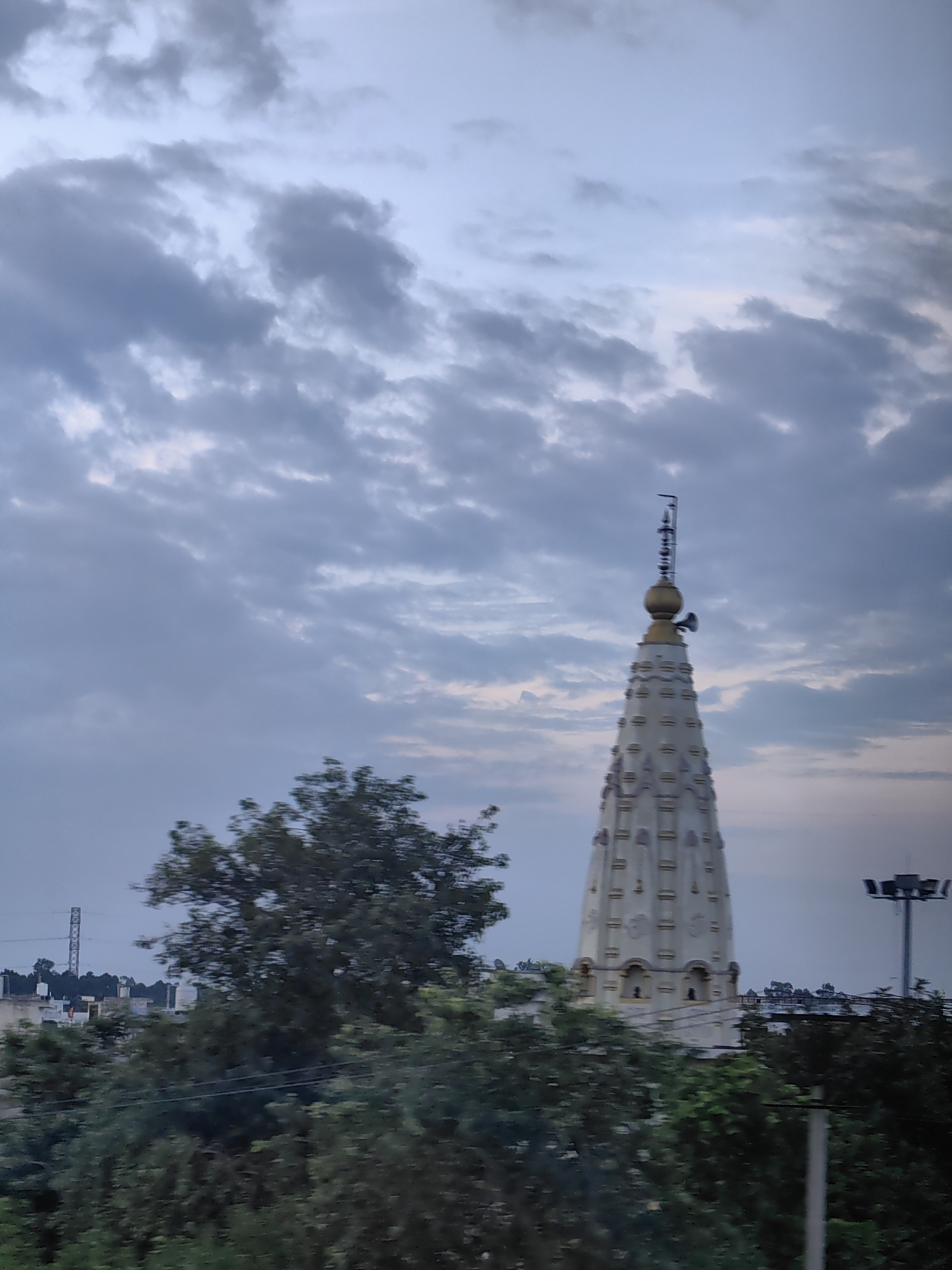 The image shows a white, tiered temple spire rising against a cloudy, pale blue sky. The spire is decorated with horizontal bands and has a golden finial topped with a small antenna. In the foreground, lush green trees obscure the lower portion of the temple and the surrounding landscape, which includes hints of buildings and a tall metal tower in the distance. The overall lighting suggests it is either early morning or late evening. This scene is located in Uchana, India.
