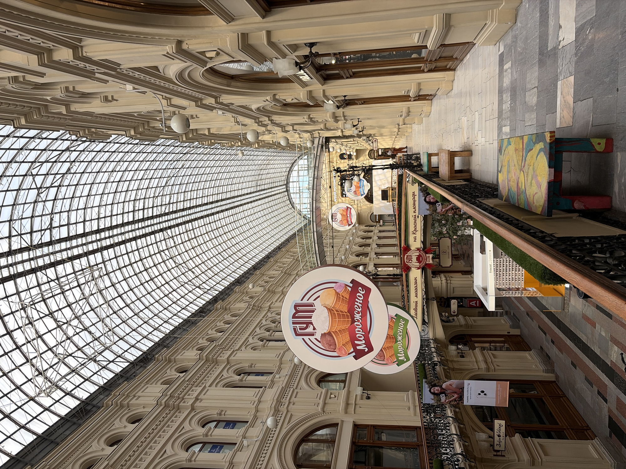 This image shows the interior of the GUM department store in Moscow, Russia, a historic shopping arcade with a distinctive glass-paneled arched roof. The perspective is from an upper level walkway, looking down the length of the building. Ornate cream-colored architectural details line both sides, with arched windows and decorative moldings.
Several round signs are suspended from the ceiling, featuring images of ice cream cones and the Cyrillic word "Мороженое" (Morozhenoye), meaning "ice cream." One sign also displays the "ГУМ" logo. Along the walkway, there are several wooden benches, one of which is brightly painted with a colorful floral pattern. People are visible in the distance, walking along the corridor. The flooring is a mix of stone tiles in various shades of grey and beige. Banners advertising products, including a Huawei phone and a brand called "Bofé Liellgi," are displayed on the railings and walls. The overall atmosphere suggests a busy, well-maintained retail environment with a blend of historical architecture and modern commerce.