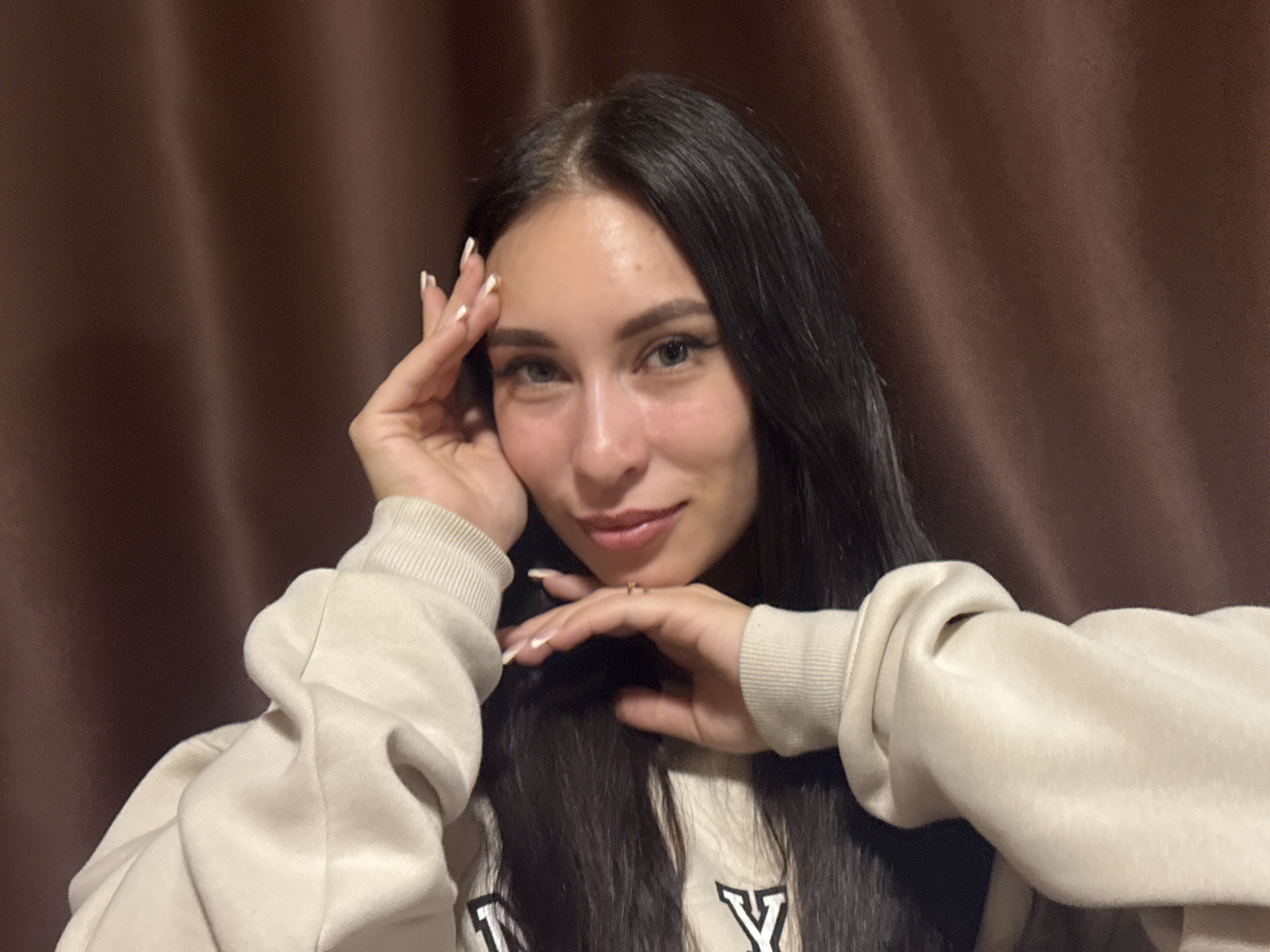 A young woman with dark hair and light skin poses indoors against a brown, textured backdrop. She wears a light-colored sweatshirt with dark lettering on the front. Her hands are positioned around her face, with manicured nails visible. The lighting appears soft and diffused, creating minimal shadows. No specific details within the image indicate that it was taken in the city of Chaikovsky, Russia. Therefore, the location cannot be conclusively determined from the photograph alone.