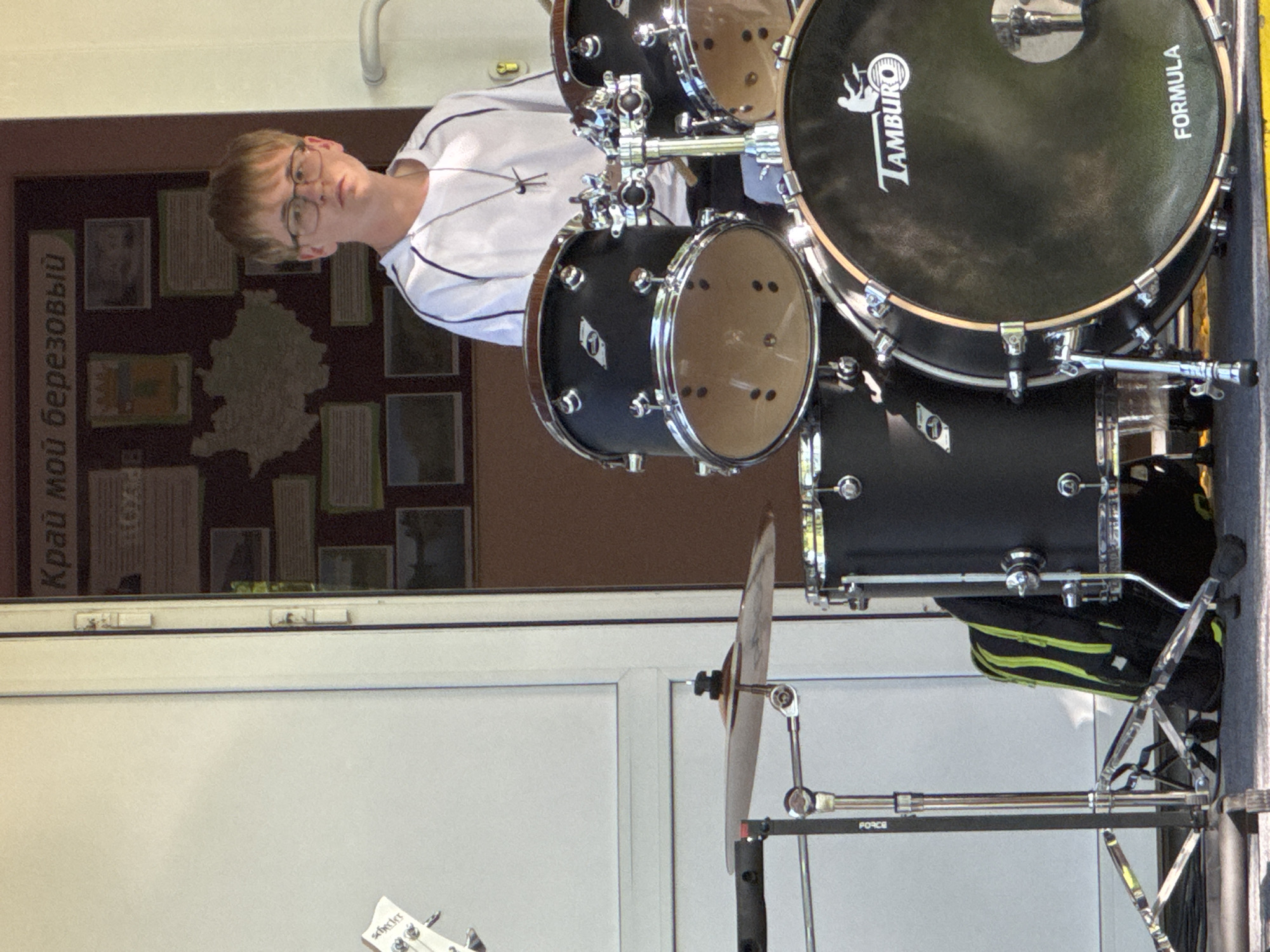 The image depicts a young man with short brown hair and glasses standing behind a drum set. He is wearing a white shirt with dark stripes on the sleeves. The drum set is black and includes a bass drum with the word "Tamburo" and "Formula" printed on it, a snare drum, and other drums. A cymbal and its stand are also visible. To the left of the drums and the man, there is a wall with a sign in Russian that begins with "Край мой березовый," which translates to "My birch land" or "My Berezovsky region." The sign is likely a display showcasing the history or geography of the Berezovsky area in Russia. Below the wall, there is a white door or wall. A portion of a white guitar is also visible at the bottom left corner of the image. Based on the Russian text "Край мой березовый" visible in the image, it's likely the location is in or near Berezovsky, Russia.