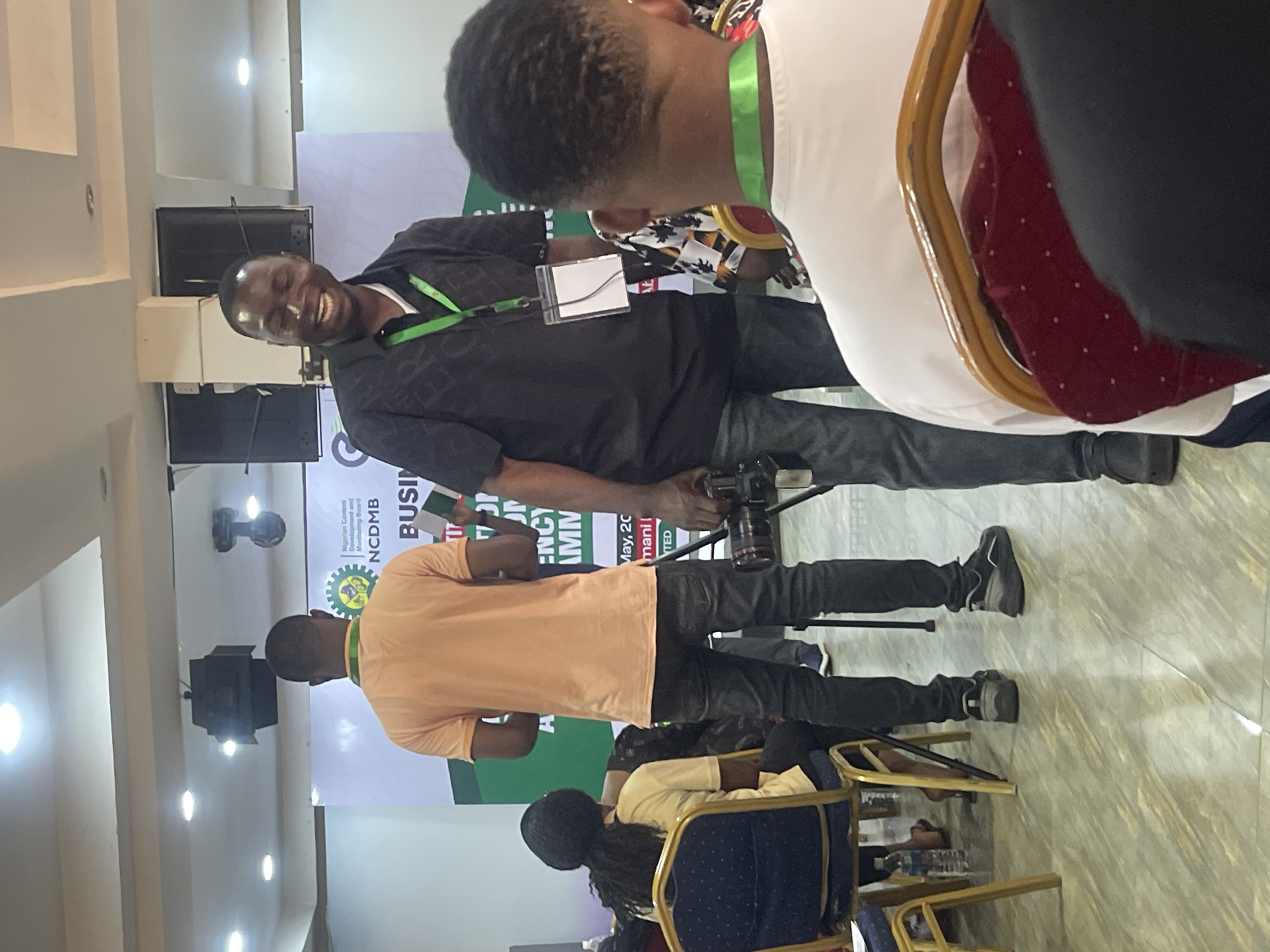 The image was taken at Galadimawa Village in Nigeria, and features a gathering of people, possibly at a conference or business event. A man in a black shirt, jeans, and sneakers smiles while holding a camera. He is wearing a lanyard with a badge, partially obscured. Another man in a peach-colored shirt stands in front of the backdrop bearing the logo "NCDMB" (Nigerian Content Development and Monitoring Board), indicating the event’s likely focus. The backdrop includes the words "BUSINESS" and "ENERGY," suggesting the event theme. Several people are seated on chairs, and the scene is lit by overhead lighting. The environment appears to be an indoor venue with pale-colored walls and a visible ceiling structure.