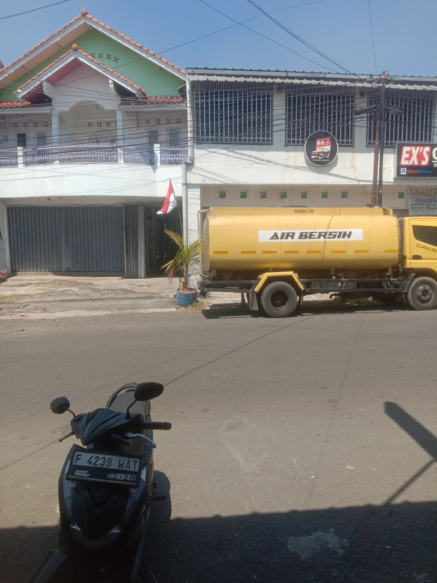 The image depicts a street scene in Cianjur, Indonesia, on a sunny day. In the foreground, a black motorcycle is parked facing away from the camera, its license plate reading "F 4239 WAT" and a sticker indicating "02.28". Behind the motorcycle, a yellow water truck with "AIR BERSIH" (Clean Water) printed on its side and "8000LTR" indicating its capacity is parked by the roadside. The truck is parked in front of a white building with a corrugated metal garage door. To the left of the garage door, a small potted palm tree stands. Further down the street, a multi-story building with balconies and barred windows is visible. A small Indonesian flag is displayed on the balcony of the first floor. A sign with "EX'S GARAGE" is also visible on the building. The environment appears to be urban, with a paved road and buildings lining the street. The sky is clear and blue.