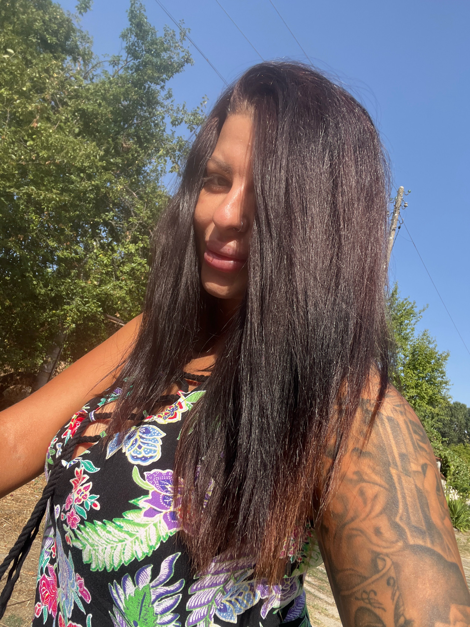 A woman with long dark hair, wearing a black dress with a floral print and a nose ring, poses for a selfie outdoors. Her left arm, visible on the right side of the frame, is covered in tattoos. Behind her, green trees and a bright blue sky are visible. The scene appears to be in Zelenikovo, Bulgaria, with a telephone pole visible in the background. The lighting suggests it is daytime, possibly late morning or early afternoon.