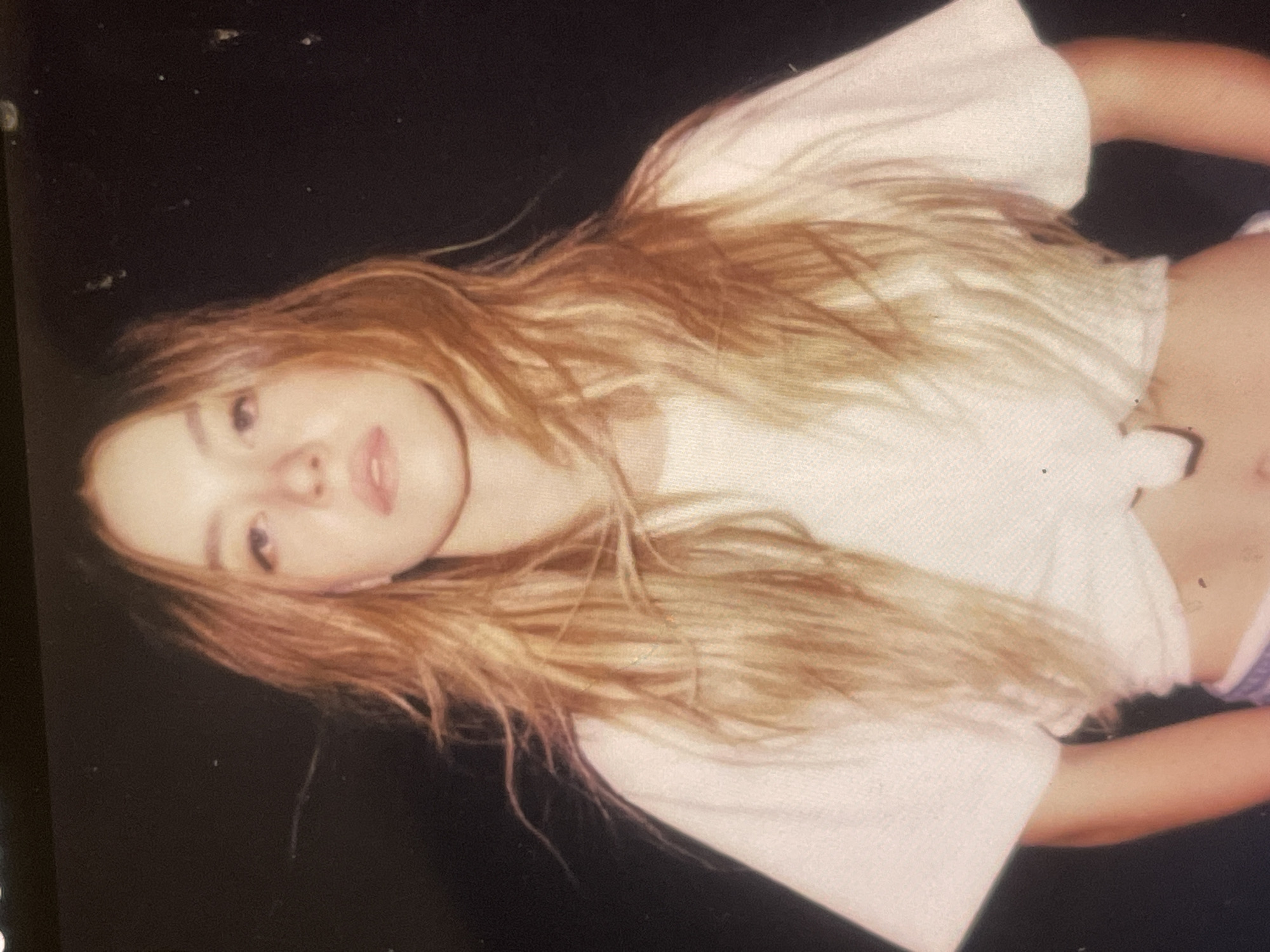 The media file displays an informal portrait of a young woman with long, flowing blonde hair, rotated 90 degrees clockwise. Her head is positioned on the left side of the frame, angled upwards and slightly to her right, with her body extending towards the right. She has fair skin, light-colored eyes, and her lips are slightly parted, showing a subtle expression that could be serene or slightly suggestive.
She is wearing a white, short-sleeved cropped top that is gathered or tied at the front, revealing her midriff. A portion of light-colored, possibly purple or striped, underwear or bottoms is visible at the lower right edge of the frame. Her arms are bent, with one arm visible at the top right of the image (as rotated).
The background is a solid, dark color, appearing black or very dark brown, suggesting an indoor setting or a plain backdrop. There are a few small, light specks scattered across the dark background, which may be dust or photographic artifacts. The lighting appears soft and illuminates her face and hair.
The image evokes a casual and relaxed mood. There are no specific location cues visible within the image itself, though the provided context indicates Poltava, Ukraine. No text is visible in the media file.