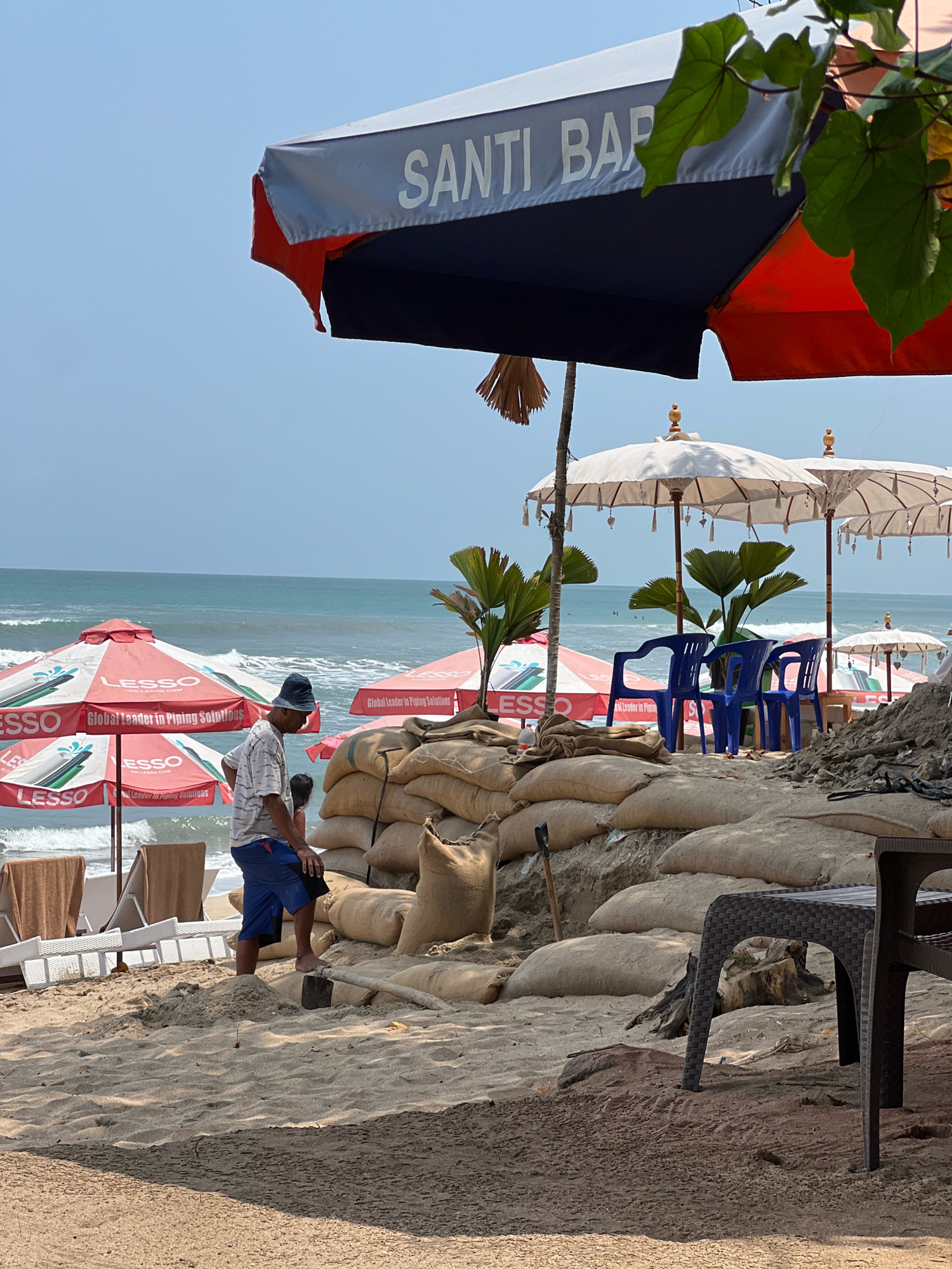 A man wearing a hat and shorts stands on a beach in Kuta, Indonesia, near a large pile of sandbags. He appears to be working with a shovel and some sand. In the background, the ocean is visible with small waves and a few surfers in the water. Several beach umbrellas are set up, some with red and white branding. There are also some blue plastic chairs and lounge chairs on the beach. A table and chairs are also visible in the foreground. The sky is clear and blue. The overall scene suggests a beach setting that may be undergoing some sort of construction or flood preparation.