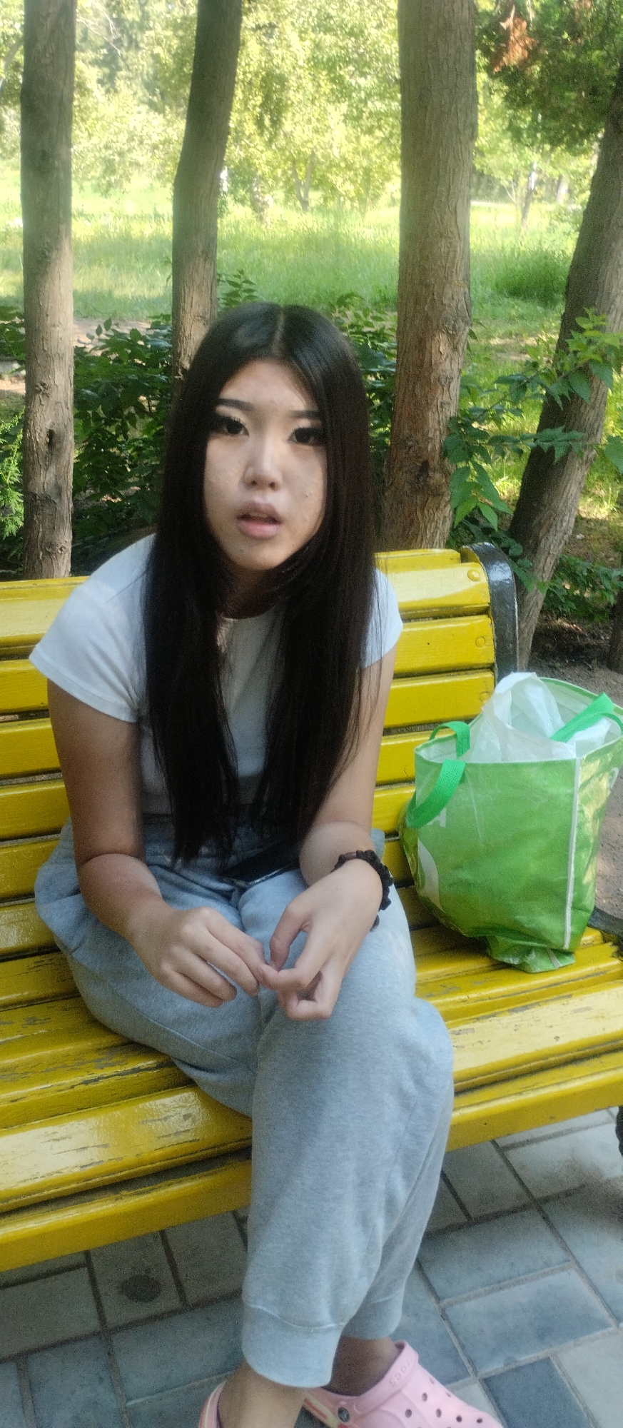 A young East Asian female with long black hair and heavy eye makeup sits on a bright yellow park bench, looking towards the camera with a neutral expression. She is wearing a simple white t-shirt and grey sweatpants, and pink Croc-style shoes are visible on her feet. Her hands are clasped loosely in her lap, and she is wearing a dark beaded bracelet on her left wrist. To her right on the bench is a green reusable shopping bag, partially filled with white plastic bags. The bench is situated in an outdoor setting, with trees and green foliage forming the background. The ground beneath the bench appears to be paved with square tiles. The overall impression is a casual moment captured in a park environment.
