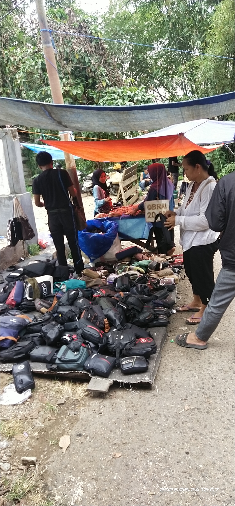 In Tamiang Layang, Indonesia, an outdoor market stall displays a large assortment of black bags, including backpacks and messenger bags, piled on a wooden platform covered with a tarp. A blue tarp is visible overhead, supported by a wooden pole and a makeshift concrete structure. Several people are present: a young man in a black t-shirt and black pants stands near the stall, a woman in a light blue hijab and patterned dress is interacting with a vendor, and another woman in a purple hijab is attending to the stall. A woman in a white shirt is also standing near the stall, looking at the items. The ground is dirt and asphalt. The background shows dense green foliage. A portion of a motorcycle is visible behind the stall. A sign on the stall reads "OBRAL 20.000". The lower right corner of the image has a timestamp: "2026.08.24 12:07".