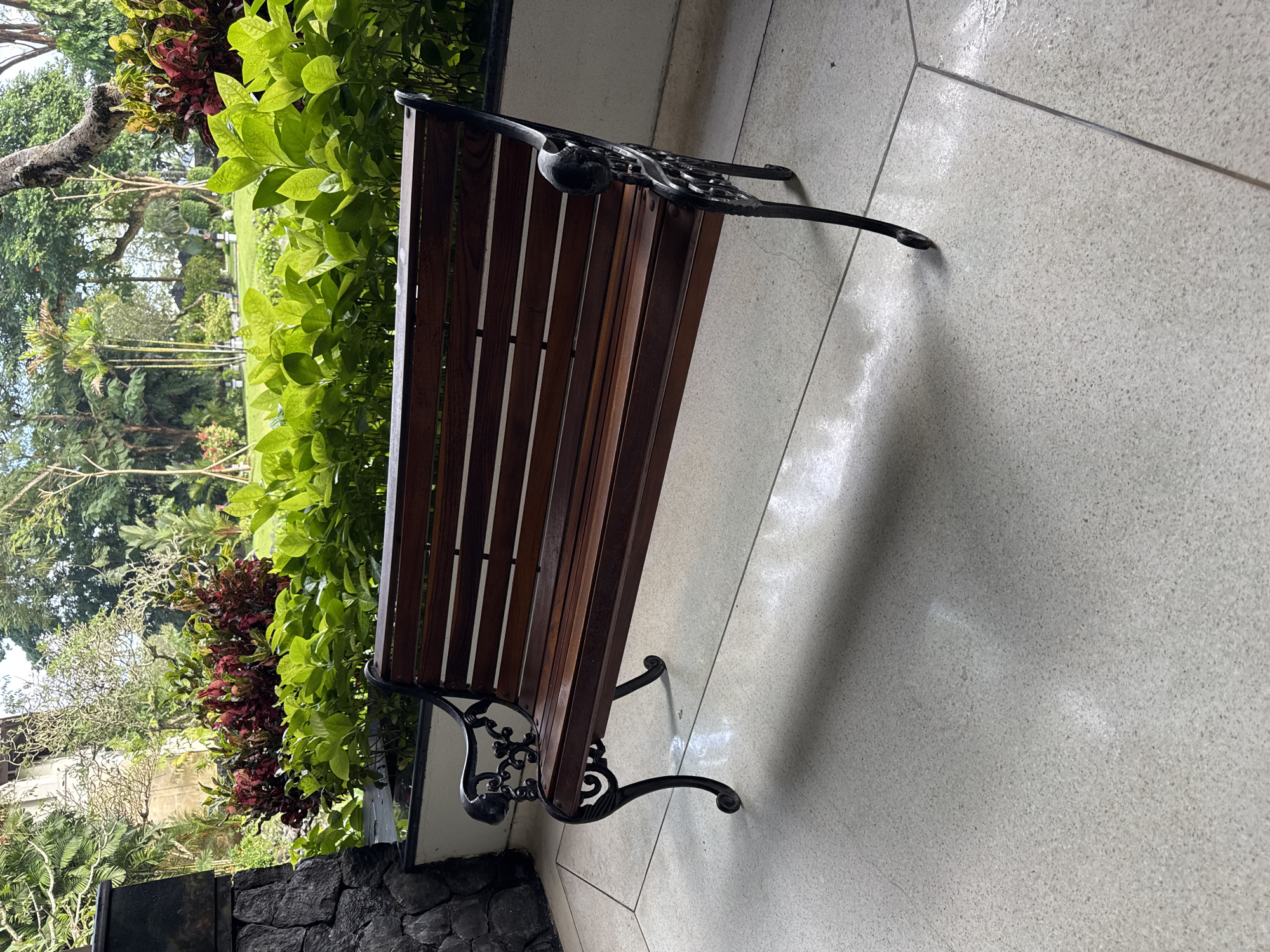 The image presents a peaceful outdoor scene in Kuta Selatan, Indonesia, centered around a park bench. The bench features dark wooden slats for its seat and backrest, complemented by ornate black wrought iron legs and armrests with intricate, scrolling designs.
The bench rests on a light-colored, speckled tiled floor, which shows visible grout lines and areas where sunlight reflects brightly, indicating it is daytime with clear or partly cloudy weather. Soft shadows are cast from the bench onto the floor.
To the left of the bench, a vibrant arrangement of tropical foliage forms a lush boundary. Bright green, densely leafed bushes dominate the foreground, interspersed with darker red and maroon leafy plants, possibly crotons. Beyond this immediate greenery, a landscaped garden or park area unfolds, featuring well-maintained grass and various mature trees, including a visible gnarled trunk and hints of palm trees in the background, characteristic of a tropical environment. A low, dark stone wall or planter box is visible in the bottom left.
No people are present, and there is no visible text. The scene conveys a sense of tranquility and an inviting atmosphere within a well-tended tropical garden setting.