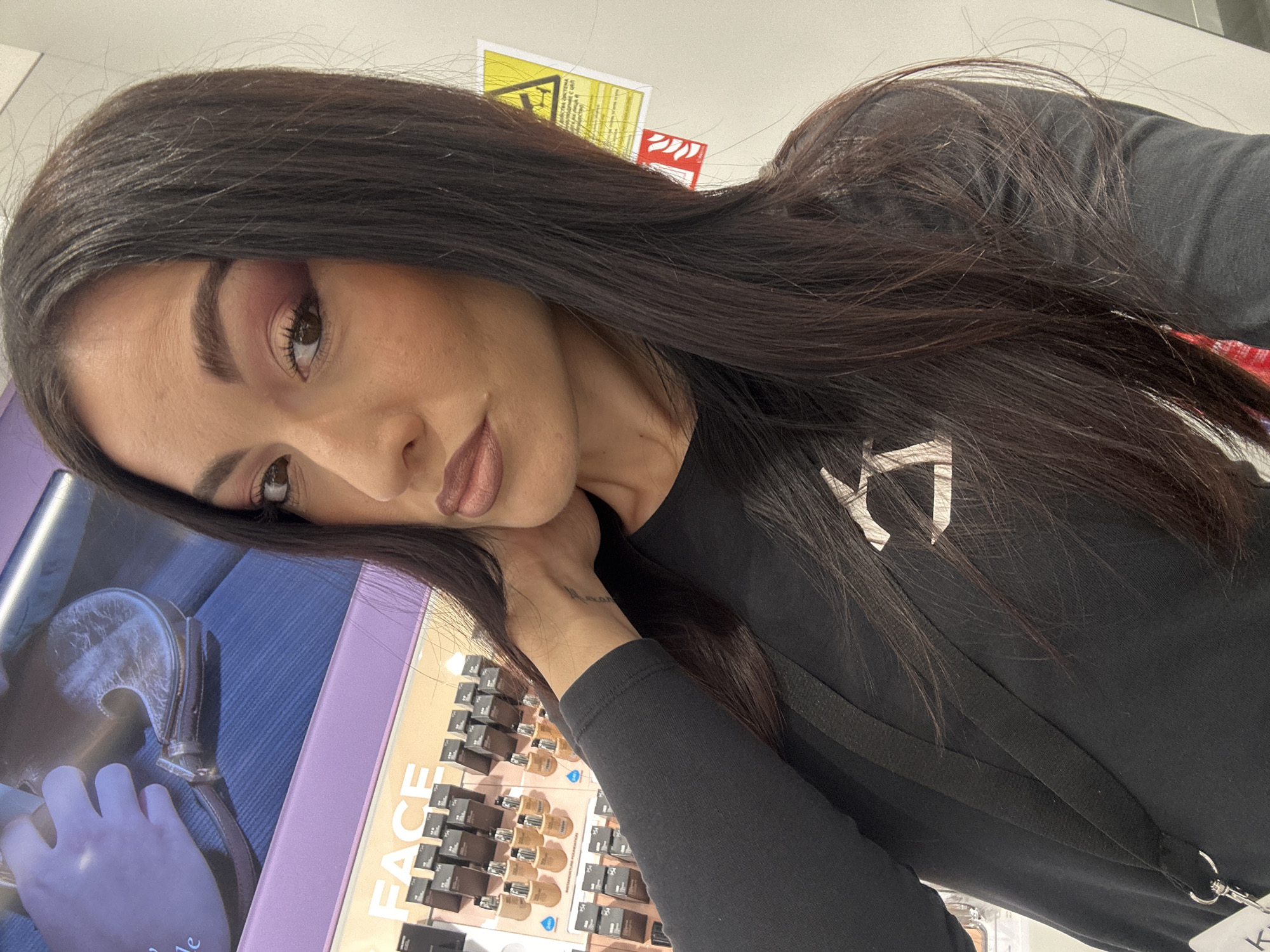 The image features a young woman with long, dark brown hair, dark eyebrows, and a thoughtful expression, posing for a selfie or close-up photograph. She is wearing a black long-sleeved top and has a black lanyard with a metal ring visible on her chest. A small tattoo is visible on her left wrist. Her makeup includes pinkish eyeshadow, dark mascara, and a neutral-toned lipstick.
The setting appears to be an indoor commercial space, likely a beauty store or a makeup counter. In the background, there is a display shelf holding numerous small bottles or containers, characteristic of foundation or other makeup products. Part of a purple display unit is visible, along with a sign that prominently features the word "FACE" in white capital letters. Above the woman's head, parts of two warning signs are visible: one yellow with black symbols, and one red with white symbols, possibly indicating safety or fire-related information. To the far left, a portion of what looks like a magazine or book page shows an image of a hand and a dark purse.
The woman is looking directly towards the camera with her head tilted, and her left hand is gently placed near her neck/shoulder. The overall lighting suggests an indoor environment.