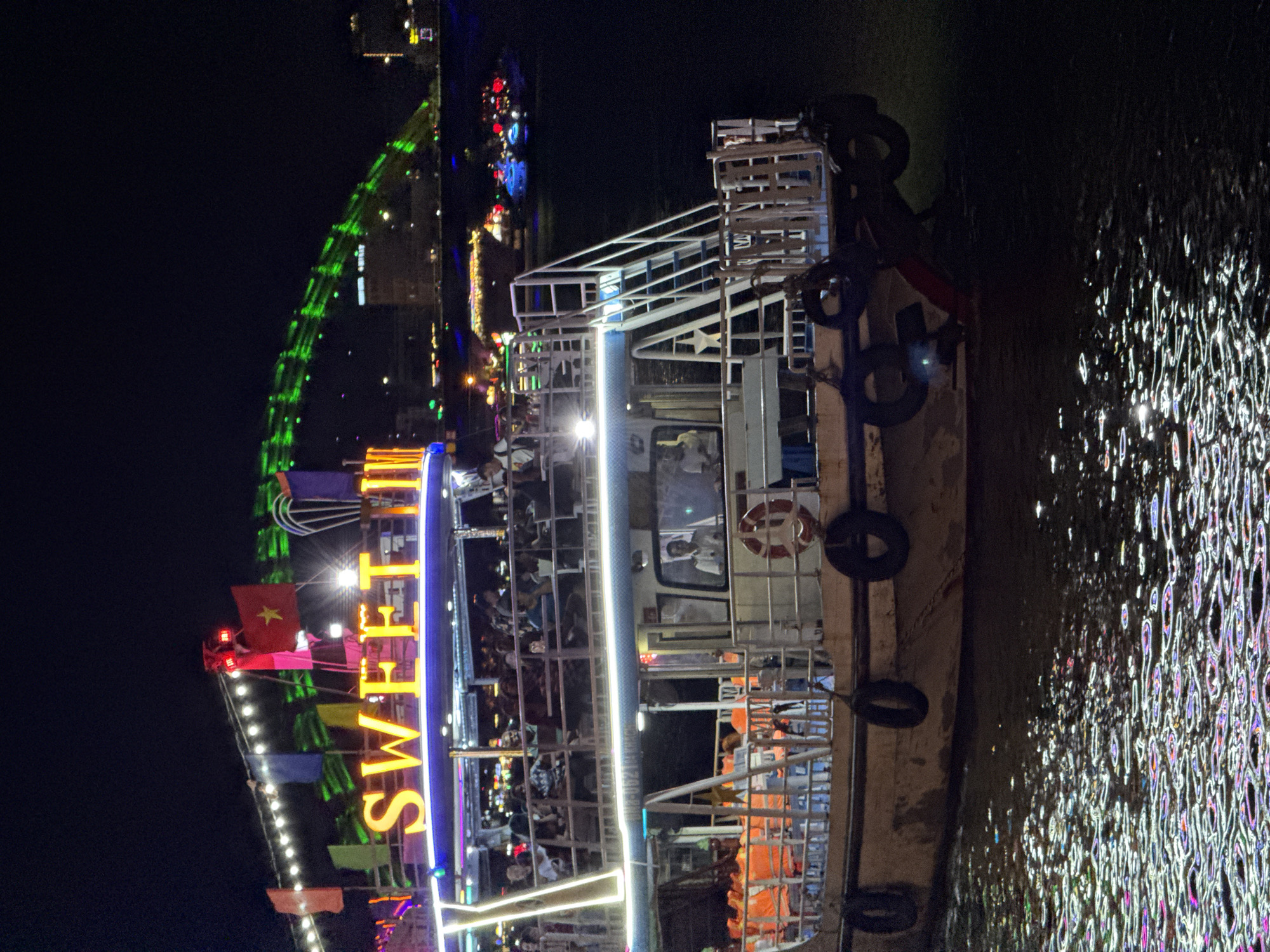 The image displays a vibrant nighttime scene featuring a large, brightly lit boat on a body of water, likely in Đà Nẵng, Vietnam. The boat is adorned with numerous colorful lights, including prominent neon letters on its side spelling "SWEET DREAMS." A red Vietnamese flag with a yellow star is visible flying from the boat's stern.
The boat has multiple decks with white railings, and several people can be seen onboard, some standing and others seated, suggesting a recreational cruise or tour. A lifebuoy is attached to the boat's side, and several black tires hang as fenders along its hull.
The dark water next to the boat reflects the myriad of lights, creating a shimmering, almost kaleidoscopic pattern of bright streaks and dots in various colors like white, blue, pink, and green. In the background, a large bridge is illuminated with distinct green lights, arching across the night sky. Beyond the bridge, more lights from city buildings are visible, indicating an urban setting. The overall atmosphere is lively and festive, typical of an evening river experience in a city.
