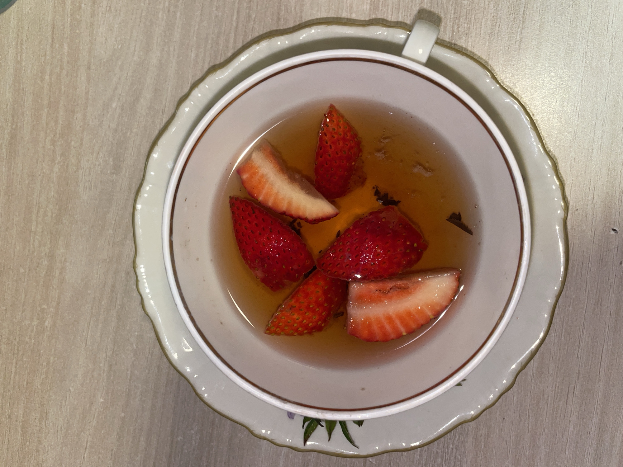 A top-down shot shows a teacup filled with light brown liquid and several slices of strawberries. The teacup sits on a decorative saucer. The background is a light brown, slightly textured surface resembling wood. The scene is indoors. The exact location and the city of Boreti, Montenegro, cannot be determined from the image. No people or actions are visible.
